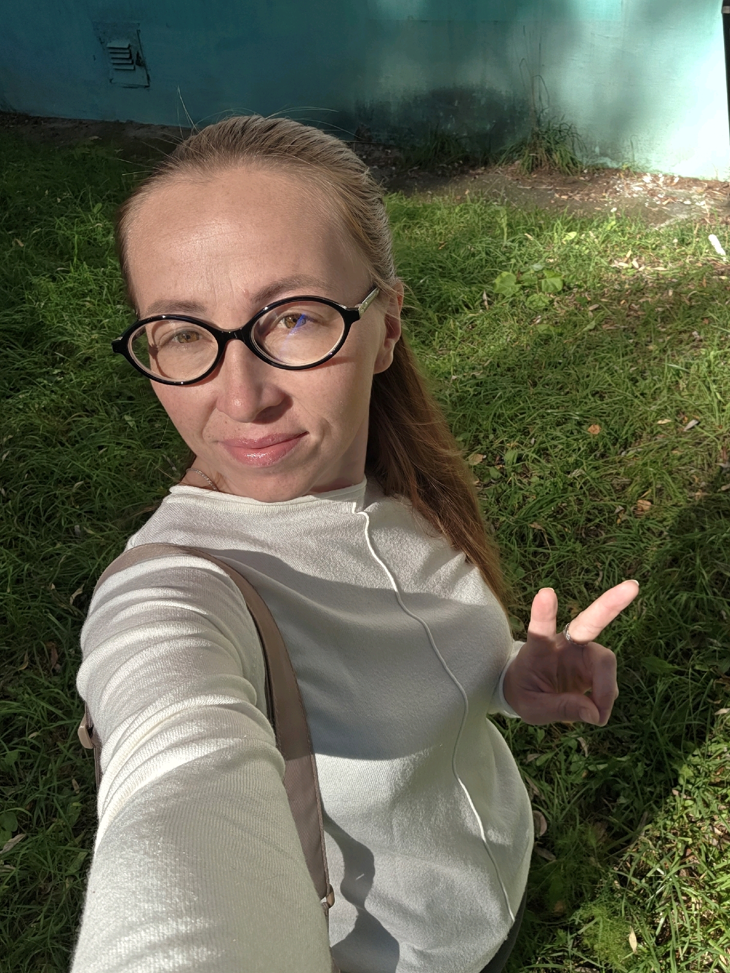 A woman with blonde hair pulled back and wearing black-rimmed glasses takes a selfie outdoors. She is wearing a cream-colored long-sleeved sweater and a light brown strap of a bag is visible across her shoulder. Her right hand is raised, and she is making a peace sign. She is standing on green grass, with a teal-colored wall behind her. The image appears to have been taken during the daytime, with shadows cast by the sun. The location is the city of Chaikovsky, Russia.