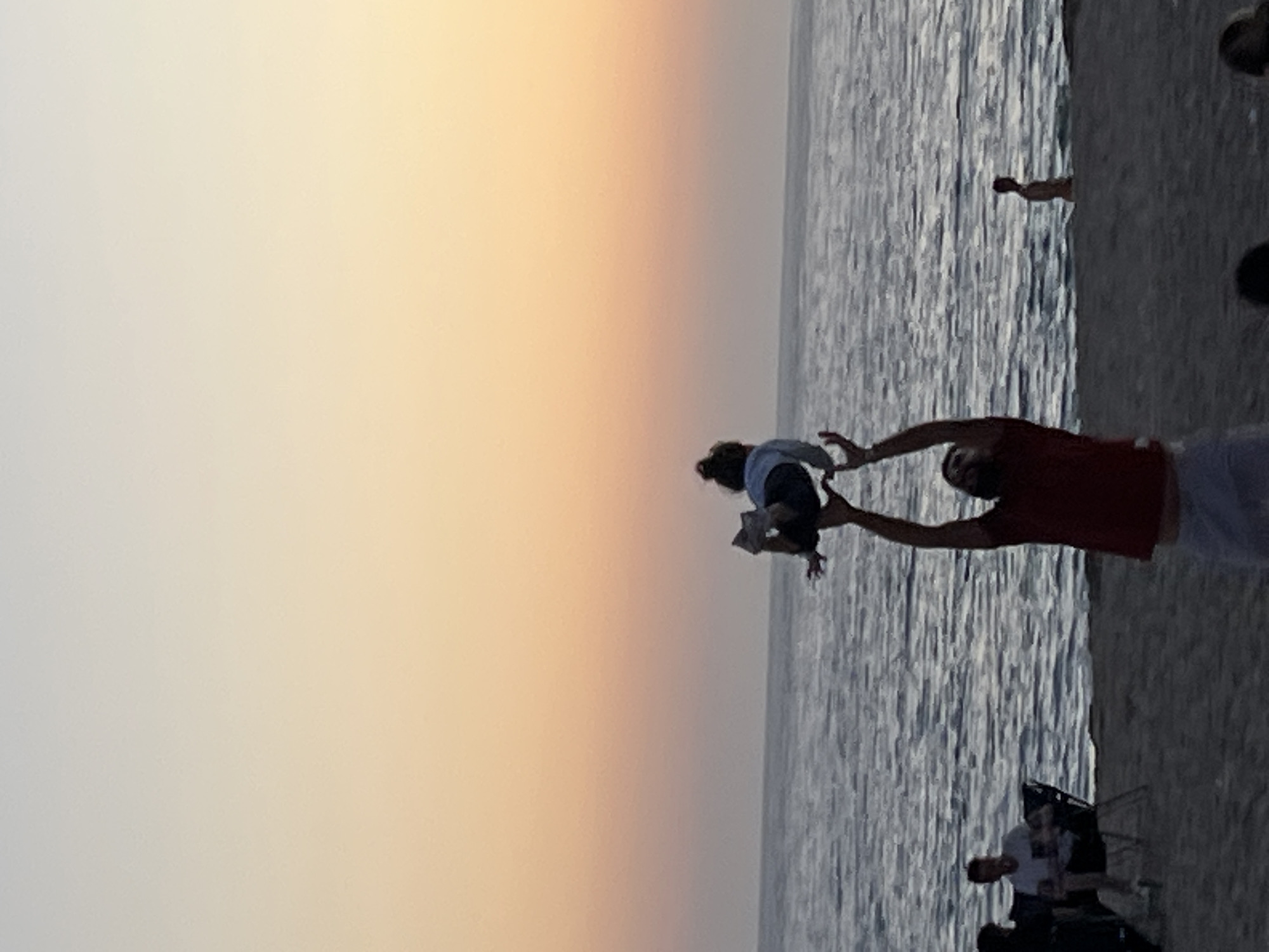 The image captures a scene at a beach in Alanya, Turkey, during sunset. A man in a red shirt and white shorts is lifting a child into the air, both facing the ocean. The water extends behind them, stretching toward the horizon where the sky transitions from a light orange near the water to a faded white towards the top of the frame. Several other people are visible in the background, with some appearing to be sitting in chairs and others standing. The beach itself, consisting of sand, is visible along the right edge of the frame.