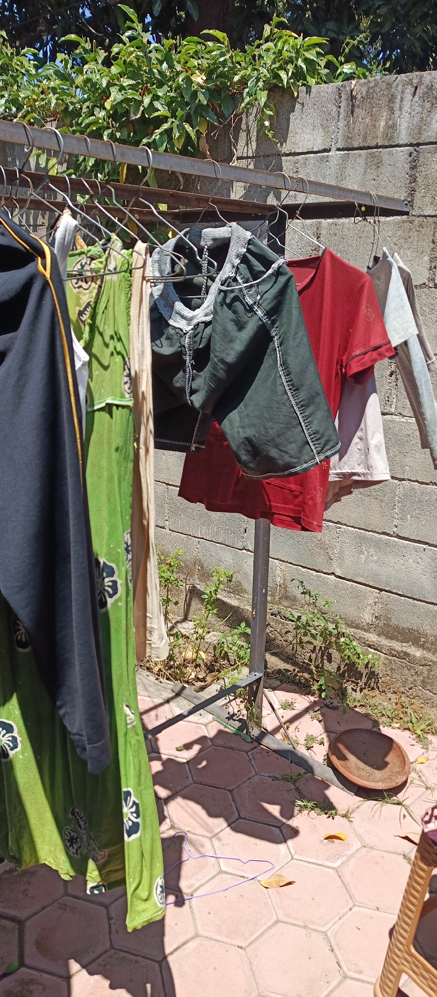 This image depicts a clothesline with several garments hanging to dry in Arjasa, Indonesia. A metal drying rack supports multiple items of clothing, including a black hooded jacket with a yellow zipper, a long green dress with a floral pattern, a beige garment, a dark green shirt, a red t-shirt, and a light gray t-shirt. The clothes are hung on metal hangers. Behind the clothesline is a brick wall, and above it, lush green foliage from a tree is visible. The ground beneath the clothesline is paved with hexagonal tiles and shows the dappled shadows of the hanging clothes and surrounding vegetation. A small, overturned brown clay dish is on the ground, along with a few fallen leaves and some sparse green plants growing near the wall. The overall scene suggests an outdoor domestic setting in Arjasa.