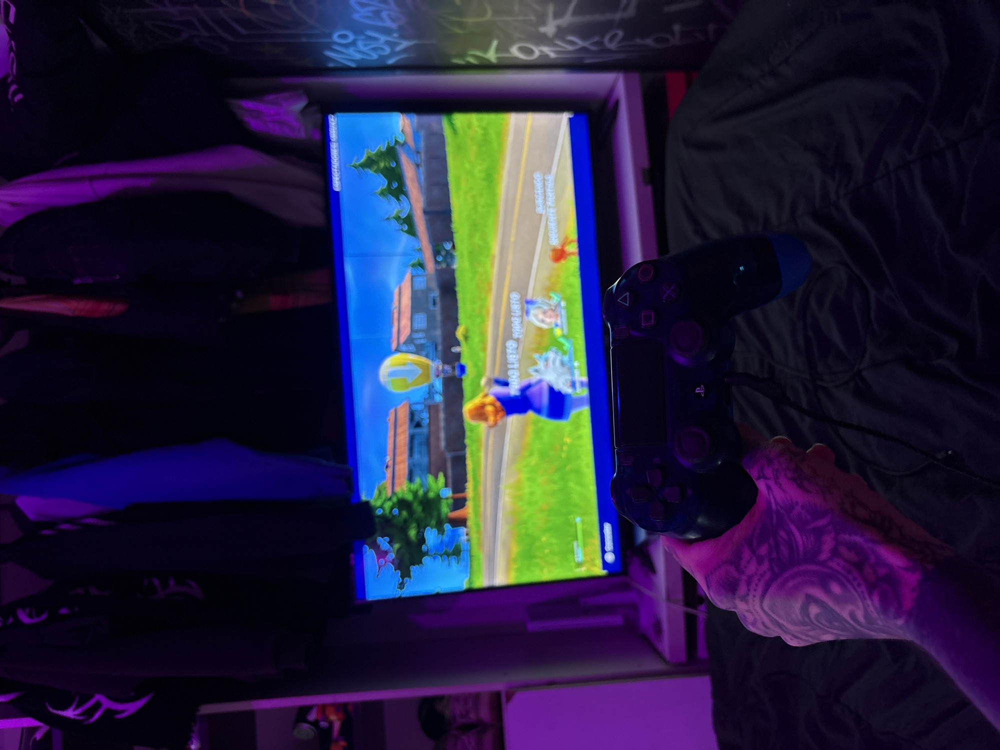 A person with a heavily tattooed right arm and hand is holding a black PlayStation 4 controller. The controller is connected by a wire to a screen displaying a game. The game is Fortnite. The screen is mounted in what appears to be a shelf or compartment. The room has clothes, bedding, and graffiti on the wall. The location cannot be determined from the provided details.