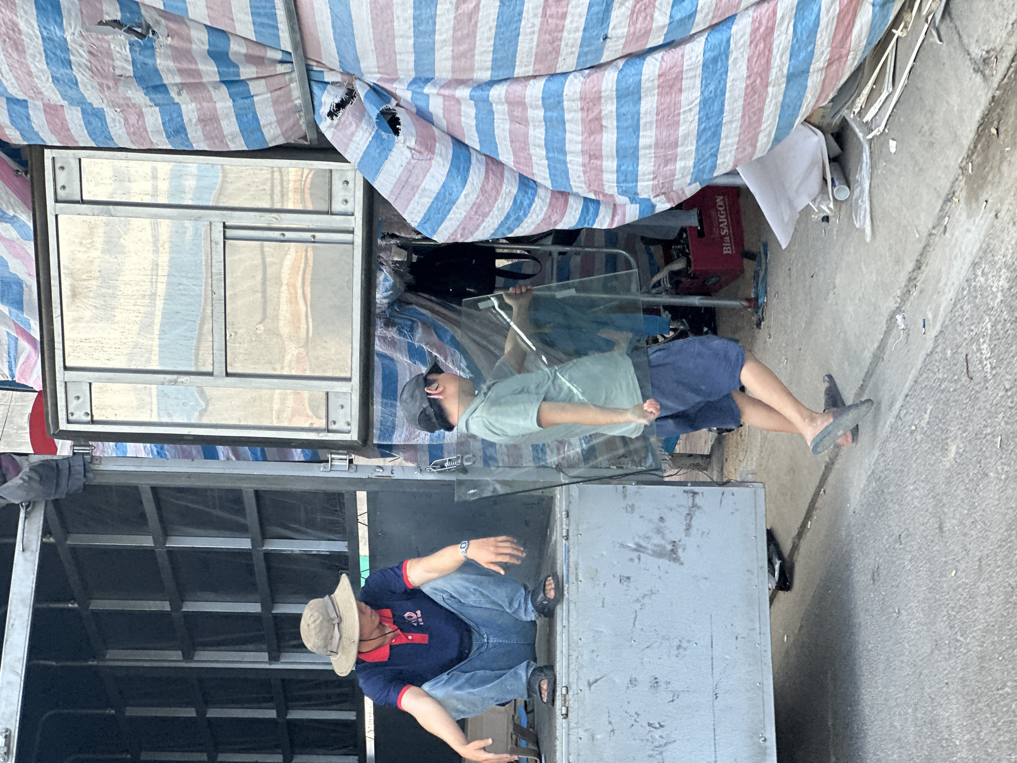A man in a straw hat and blue jeans sits on a truck bed on a street. The street is located in the city of Nha Trang Ward, Vietnam. Another man is standing and holding a glass panel. Both are working on or assembling the truck bed. There is a striped tarp overhead.