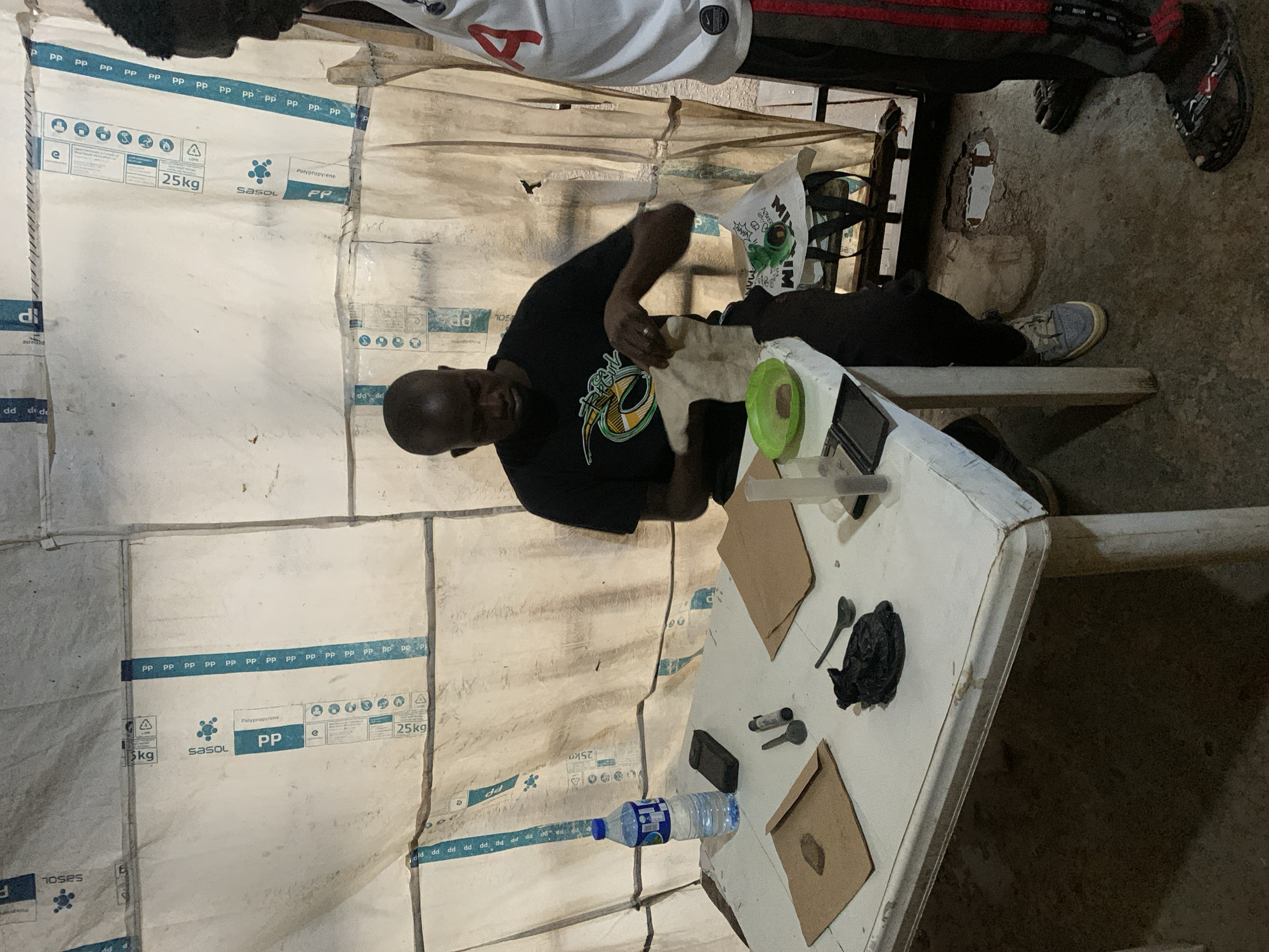 The image depicts an indoor scene with a man in a black t-shirt actively engaged with materials spread on a white table. He is holding a light-colored cloth or paper and appears to be examining or manipulating its contents. On the table, there are several items, including a bottle of water, a dark-colored phone, two small metal spoons, a small vial, a small green bowl containing a brown substance, and two pieces of brown paper, one with a stain. A black, lumpy object, possibly a bag of material, is also on the table near a spoon. The background consists of white, opaque sheeting, possibly tarpaulin, with blue striped tape running horizontally and vertically across it, displaying text like "25kg," "sasol," and "polypropylene." The floor appears to be concrete, with some unevenness and a small puddle of water visible in the lower right section of the frame. Another person, wearing a white shirt with a red number "4" and black pants with red stripes, stands to the right of the frame, partially visible. The setting suggests a makeshift or temporary structure, possibly an outdoor market stall or a temporary shelter in a developing urban environment. Without further context, it is difficult to pinpoint an exact location within Jos, Nigeria, but the overall aesthetic is consistent with informal settlements or makeshift structures found in such areas.