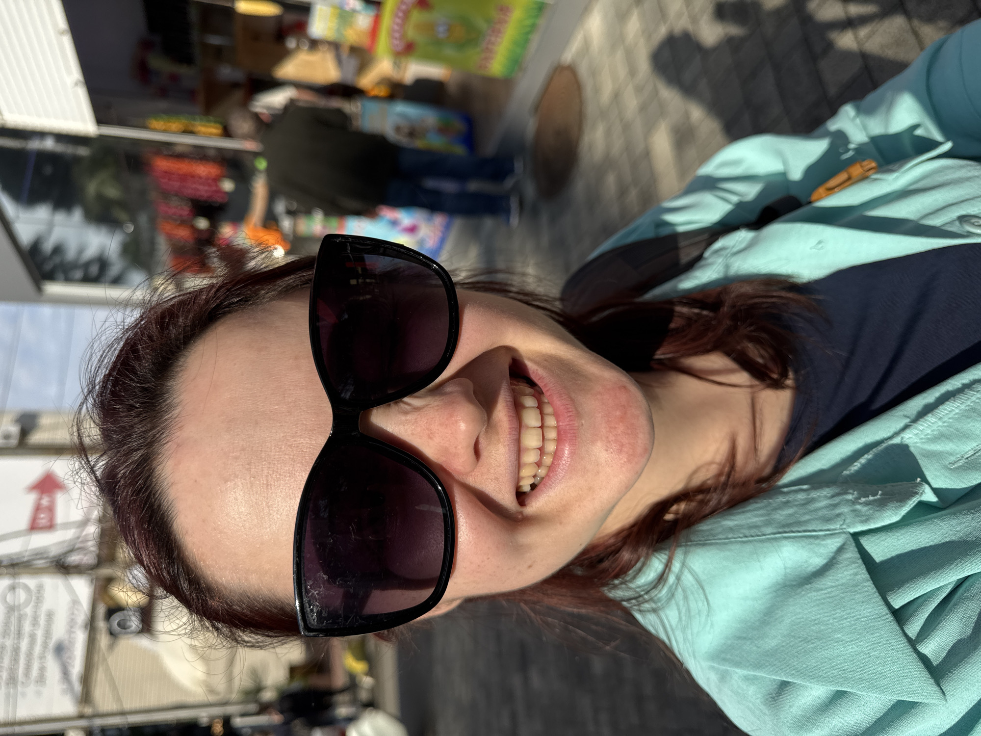 A woman is smiling at the camera, wearing dark sunglasses and a light blue collared shirt, over a dark top. She is standing outside, possibly near a shop, as reflected in the sunglasses and the background. The location is indicated by the street signs and the overall aesthetic to be Sochi, Russia.