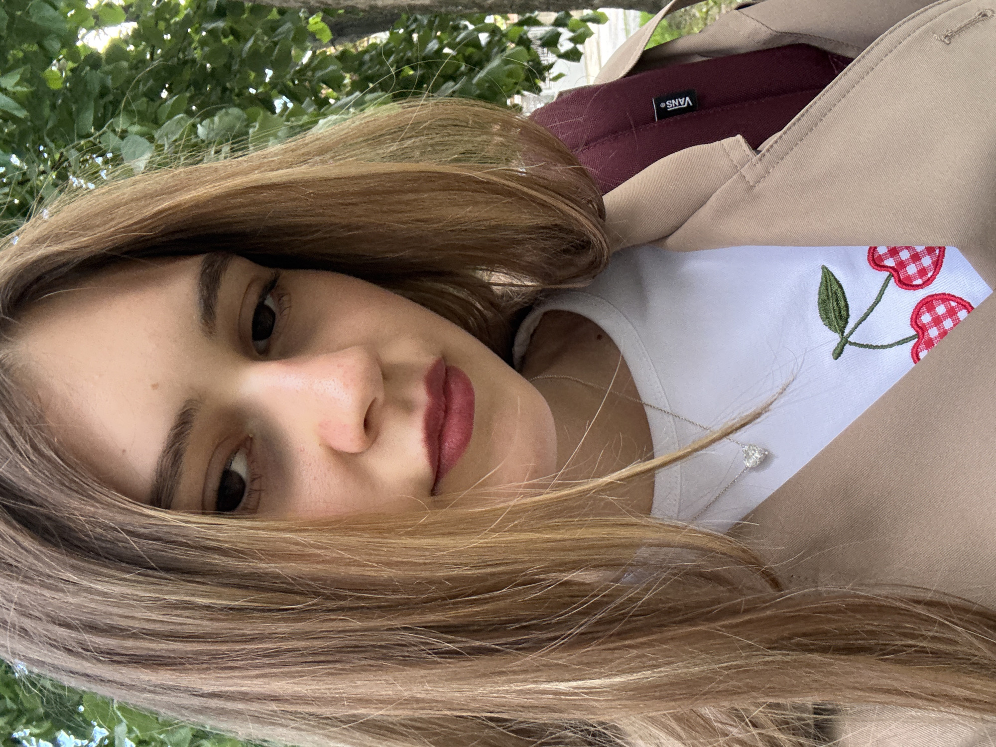 A young woman with brown hair, light skin, and dark eyes is facing the camera. She is wearing a white t-shirt with embroidered cherries and a beige trench coat. A burgundy backpack with the "Vans" logo is visible behind her shoulder. The background shows green leaves. It is not possible to determine the location within Panagyurishte, Bulgaria from this image alone.