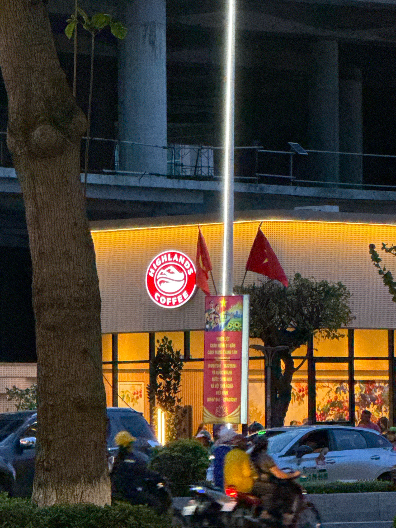 This image depicts the exterior of a Highlands Coffee shop in Nha Trang, Vietnam, at dusk. A large tree trunk dominates the left foreground, partially obscuring the storefront. The coffee shop features a bright yellow, textured facade with an illuminated "Highlands Coffee" logo in the center. Two red Vietnamese flags are visible, one on either side of the logo. A tall, bright white light fixture stands to the right of the logo, extending upwards. In front of the store, a sign with text and images is displayed. The background shows an under-construction building with concrete pillars. In the lower portion of the image, a street scene unfolds with several vehicles, including a silver car and motorcycles. People are seen on the motorcycles and near the car, indicating activity on the street. The overall lighting suggests it is either early morning or late evening.