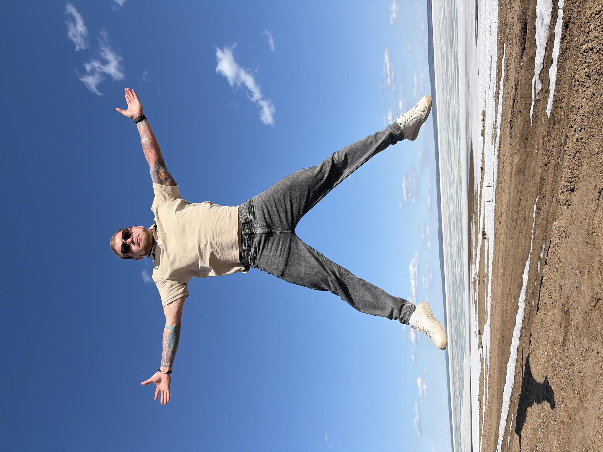 A person stands on a sandy, partially snow-covered ground with a body of water in the background, under a blue sky with scattered clouds. The person is seen from a low angle, with arms outstretched and legs spread in a "V" shape. They wear a tan shirt, gray jeans, and white sneakers, and have visible tattoos on their arms. The person is wearing sunglasses. The location is near Noviy, Russia.