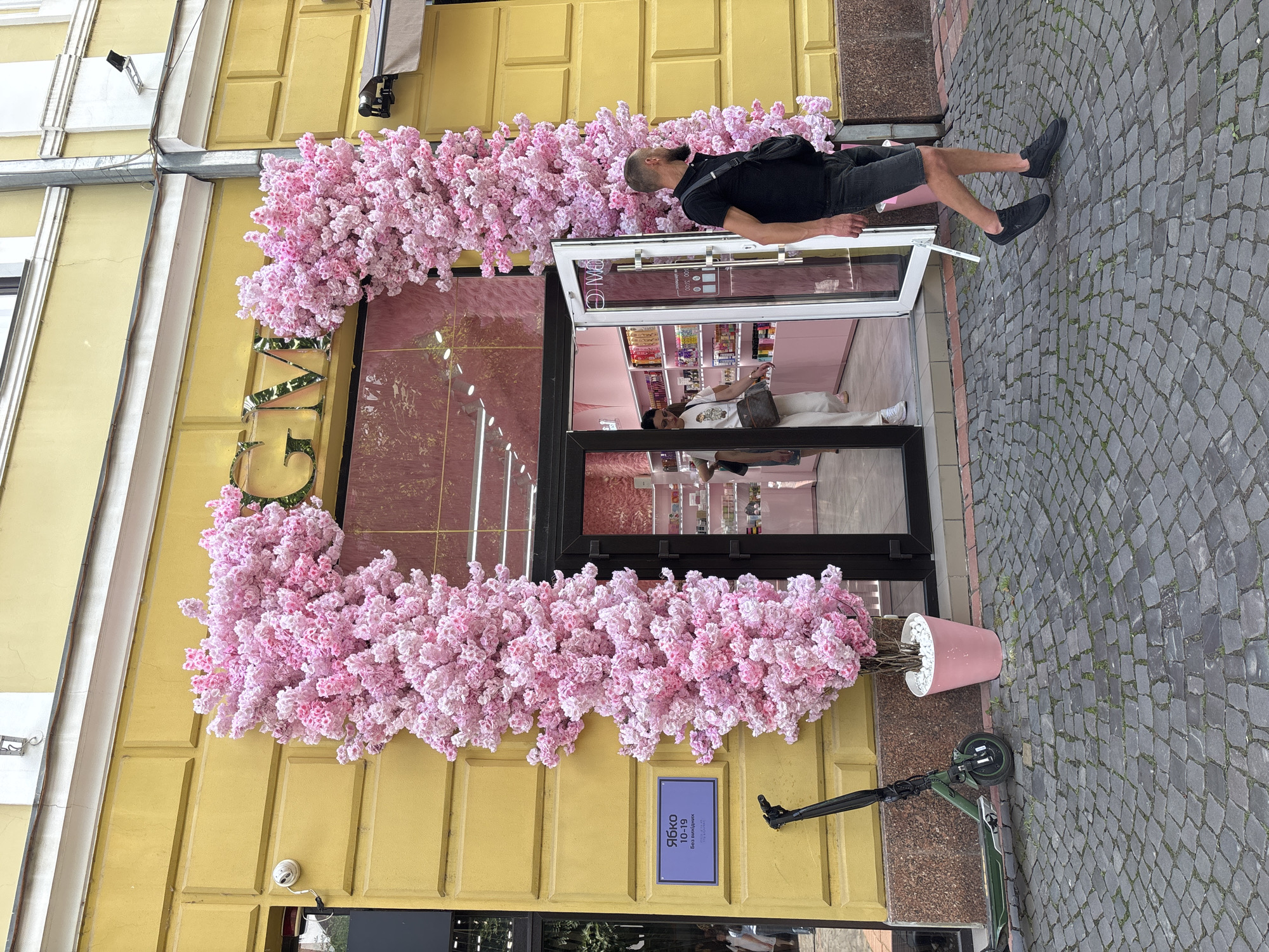 The image shows the entrance to a store decorated with artificial pink cherry blossoms. The store has a yellow facade, and the entrance is framed by the blossoms, creating an archway. Above the entrance, the store name, "GM," is visible in gold lettering. Inside the store, shelves are lined with products, and the interior is painted pink.
To the left of the entrance, a green electric scooter is parked next to a blue sign with text in Cyrillic. A man in a black t-shirt and shorts is walking away from the store entrance towards the right side of the image. Another person, partially visible, is walking into the store. The ground in front of the store is paved with cobblestones. The overall scene appears to be a street in an urban environment.