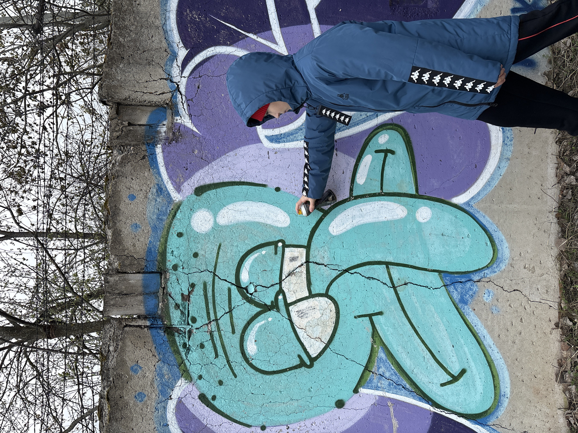 A person wearing a blue hooded jacket with black pants and a red hat is spray-painting graffiti on a concrete wall. The graffiti consists of a cartoon-style character in shades of blue and purple. The background includes bare trees and the edge of a concrete structure. The location is the city of Chaikovsky, Russia.