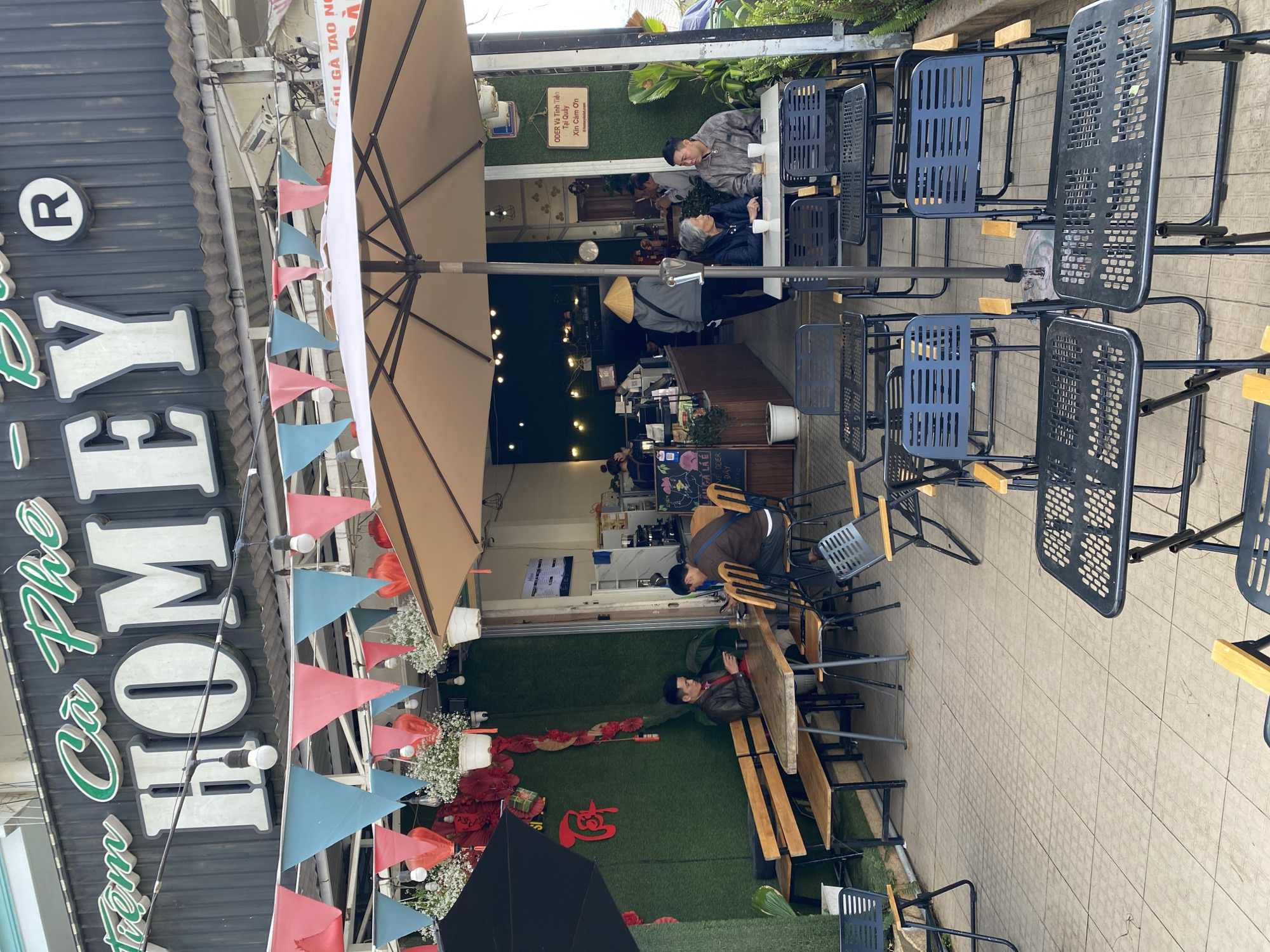 The image depicts the outdoor seating area of a coffee shop in what appears to be Lam Vien - Da Lat, Vietnam. Customers are seated at various tables, some occupied and others empty. The coffee shop is decorated with strings of triangular flags in red and blue, and there are faux grass walls with floral arrangements. A large umbrella provides shade to some of the seating area. The storefront has a sign with the name "HOMEY Cà Phê" in a stylized font. Patrons are engaging in conversations. The seating consists of metal chairs and wooden benches.