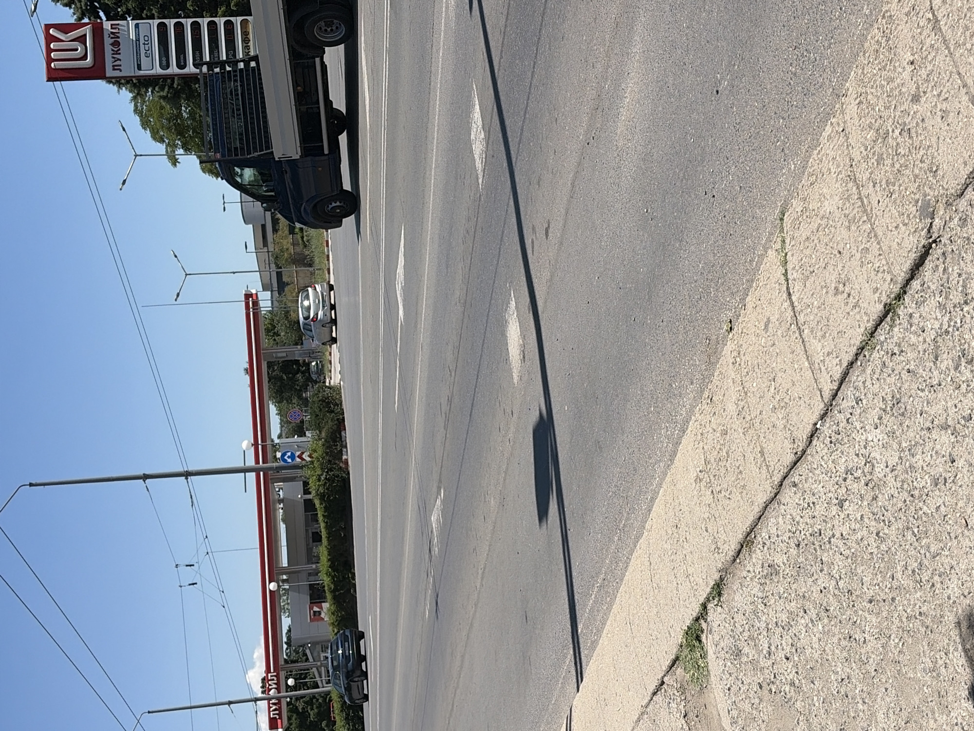 The image depicts a street scene in Stara Zagora, Bulgaria, on a clear, sunny day. In the background, a Lukoil gas station with red awnings is visible. A tall signpost for Lukoil displays fuel prices. A dark-colored flatbed truck is positioned in the right lane of the road, facing away from the camera. A silver hatchback car is in the left lane, also facing away. The road has multiple lanes marked with white lines. A concrete sidewalk runs along the left edge of the image, with some patchy vegetation growing along its edge. Overhead wires, likely for public transportation, are present in the sky.