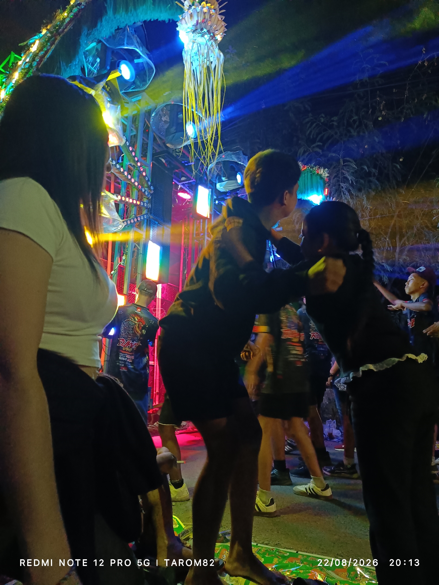 In Mundusewu, Indonesia, an outdoor night event is underway, possibly a festival or gathering. The scene is illuminated by vibrant, multi-colored lights, predominantly blue, yellow, and pink, emanating from a large, complex stage or structure adorned with numerous light bulbs, speakers, and what appears to be a jellyfish-like decoration.
In the foreground, a person with long dark hair, wearing a white t-shirt, is seated, facing away from the camera. To their right, a young man in a dark hoodie is embracing a young woman, also in dark clothing. Both are looking towards the stage.
Behind them, other individuals are visible, some wearing dark t-shirts with graphic designs, suggesting a themed event. The ground appears to be paved or a dirt surface, with some colorful mats or coverings laid out. The overall atmosphere suggests a lively and energetic event, with the lighting creating a dramatic and festive ambiance. The date and time stamp on the image indicates August 22, 2026, at 20:13.