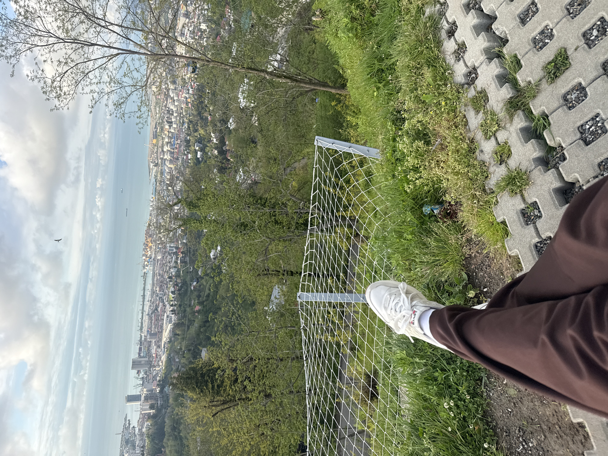 A vertically oriented image shows a view overlooking Batumi, Georgia. The viewpoint is from an elevated position with a protective fence in the foreground. The image captures a cityscape, a body of water, and a cloudy sky. A person's legs, clad in brown pants and white sneakers, are visible on the right side of the frame, close to the edge of a paved area with grass growing between the pavers. The fence consists of a white net with metal supports. The cityscape shows numerous buildings, and the body of water has ships visible. The sky is overcast.