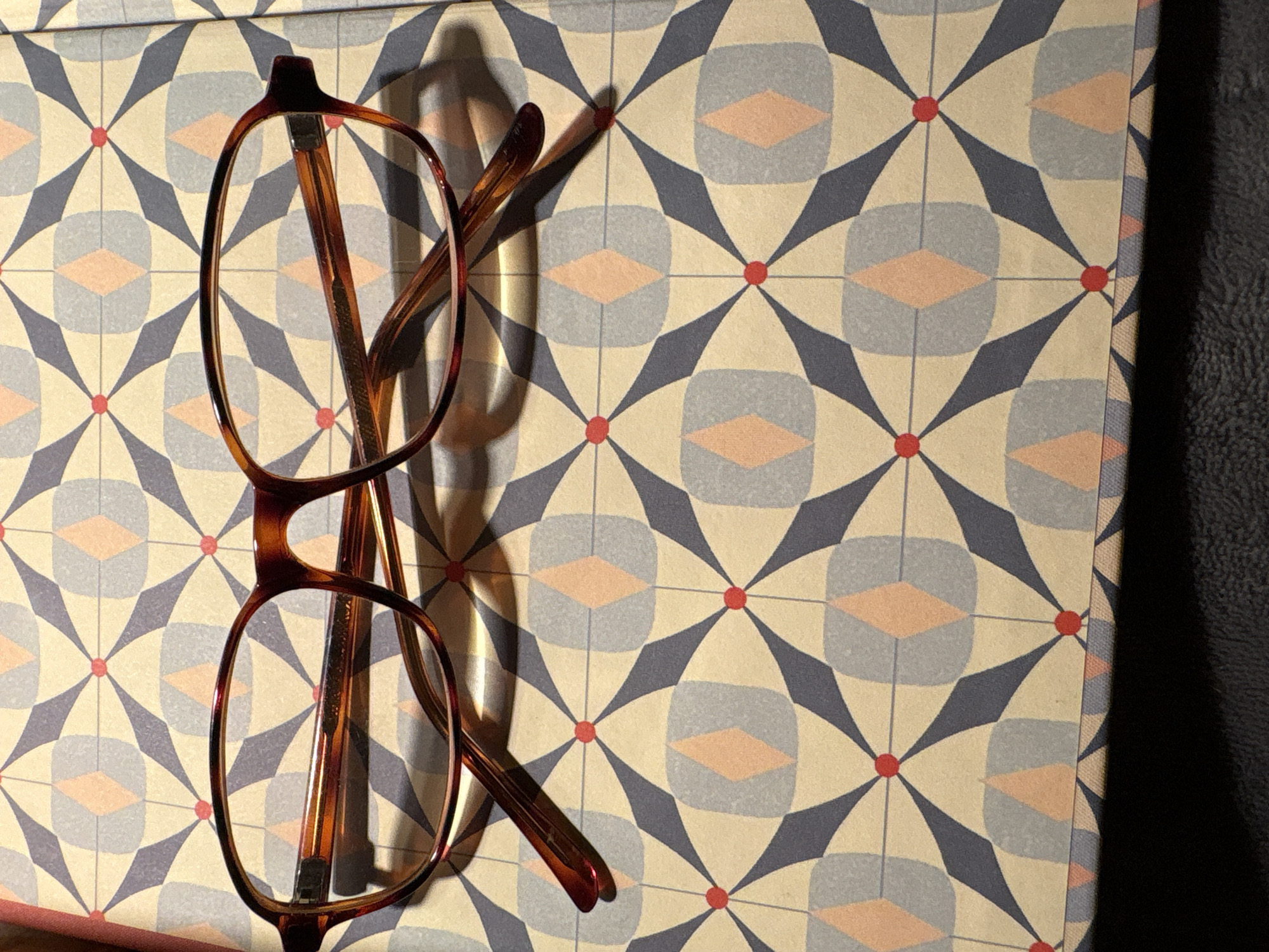 A pair of eyeglasses with tortoiseshell frames rests on a patterned surface. The pattern consists of a repeating geometric design with diamond shapes in shades of cream, gray, and coral, intersected by navy blue triangles and small red dots. The eyeglasses are positioned horizontally across the frame, with the left lens partially obscured by shadow. The right temple of the glasses is visible and curves downwards. The background appears to be a book cover or similar item with this geometric print. The lower portion of the image is dominated by a dark shadow. The location is Unknown, Unknown.