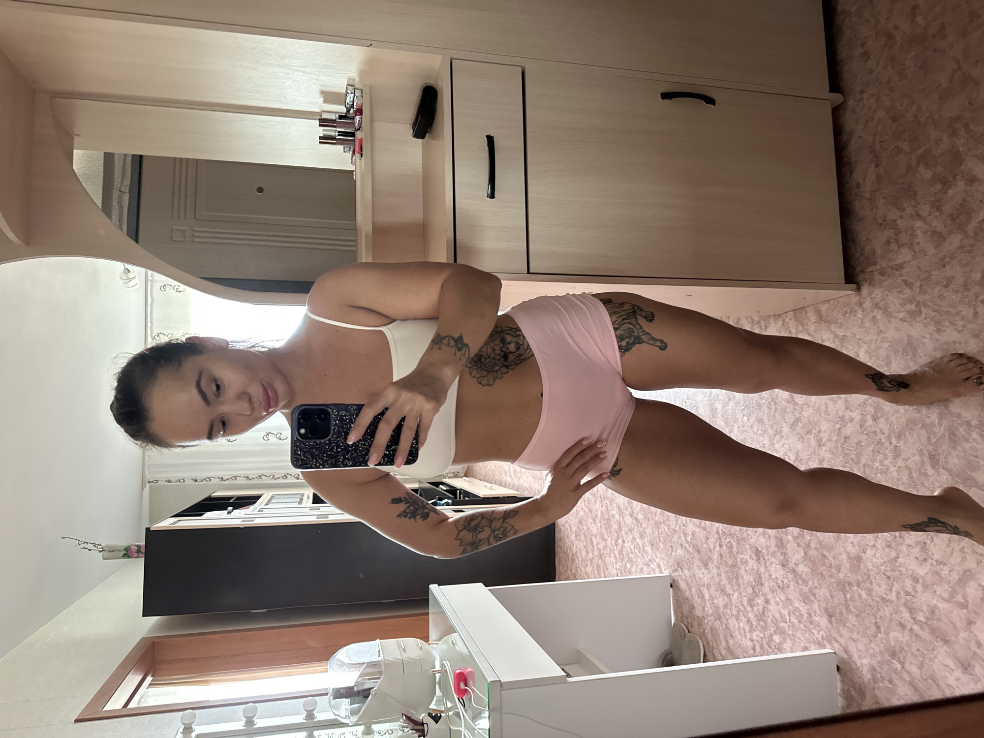 A full-body, slightly upward-angled selfie is taken by a woman with dark hair pulled back and bare shoulders, wearing a white crop top and pink shorts. She is standing in what appears to be a bedroom or dressing area. The woman has tattoos on her arms, thighs, and calves. She is holding a dark-colored smartphone in her right hand, angled to capture her reflection.
The background shows a light-colored dresser with a wooden finish, a mirror with a curved top, and a dark tall cabinet. On the dresser, several tubes of lipstick and other cosmetic items are visible. To the left, a white vanity table with a lighted mirror is partially visible, along with some makeup organizers. The floor is covered with a pinkish, marbled linoleum. The lighting appears to be natural, coming from the right side of the frame. The overall environment suggests a personal dressing space within a residential setting.
