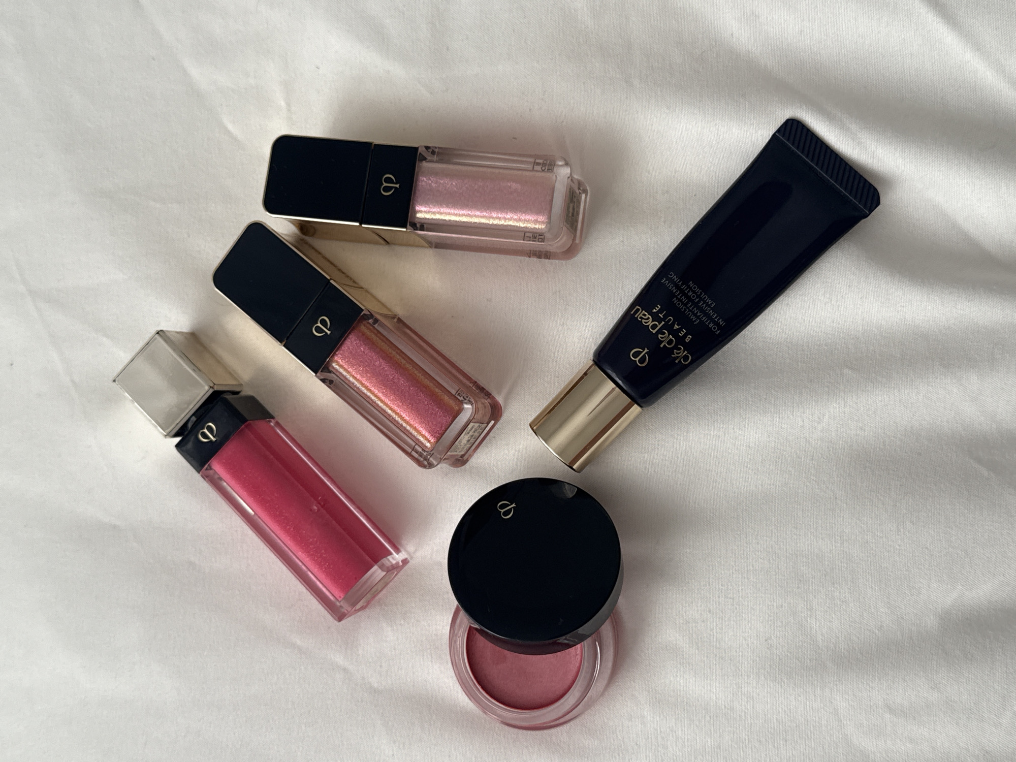 The image shows a still life arrangement of Clé de Peau Beauté makeup products on a white, textured surface, likely a bedsheet or other cloth. The items include three lip glosses in clear, rectangular containers with black and gold tops; one tube of emulsion in a dark blue container with gold accents; and one small jar of what appears to be eye or lip color with a black lid. There are no people in the image. The scene is not located in London, United Kingdom. The environment is generic, suggesting an indoor setting.