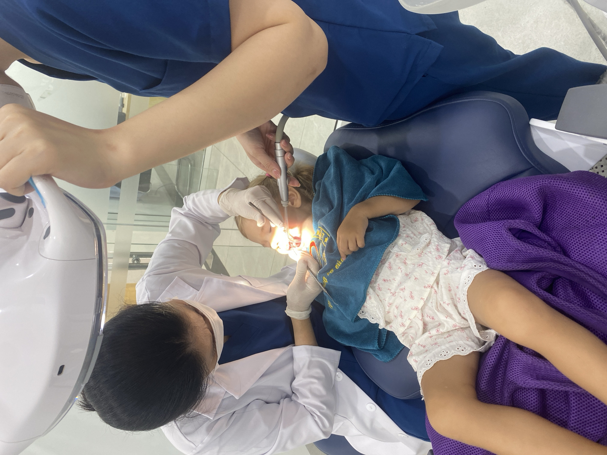 A child is lying on a dental chair, undergoing dental examination. A dentist wearing a white coat and a mask is examining the child's teeth with a dental mirror. Another person, likely a dental assistant, is holding a dental drill. The child is covered with a blue towel and wearing white shorts with floral patterns. The environment appears to be a dental clinic in Phú Thủy, Vietnam.