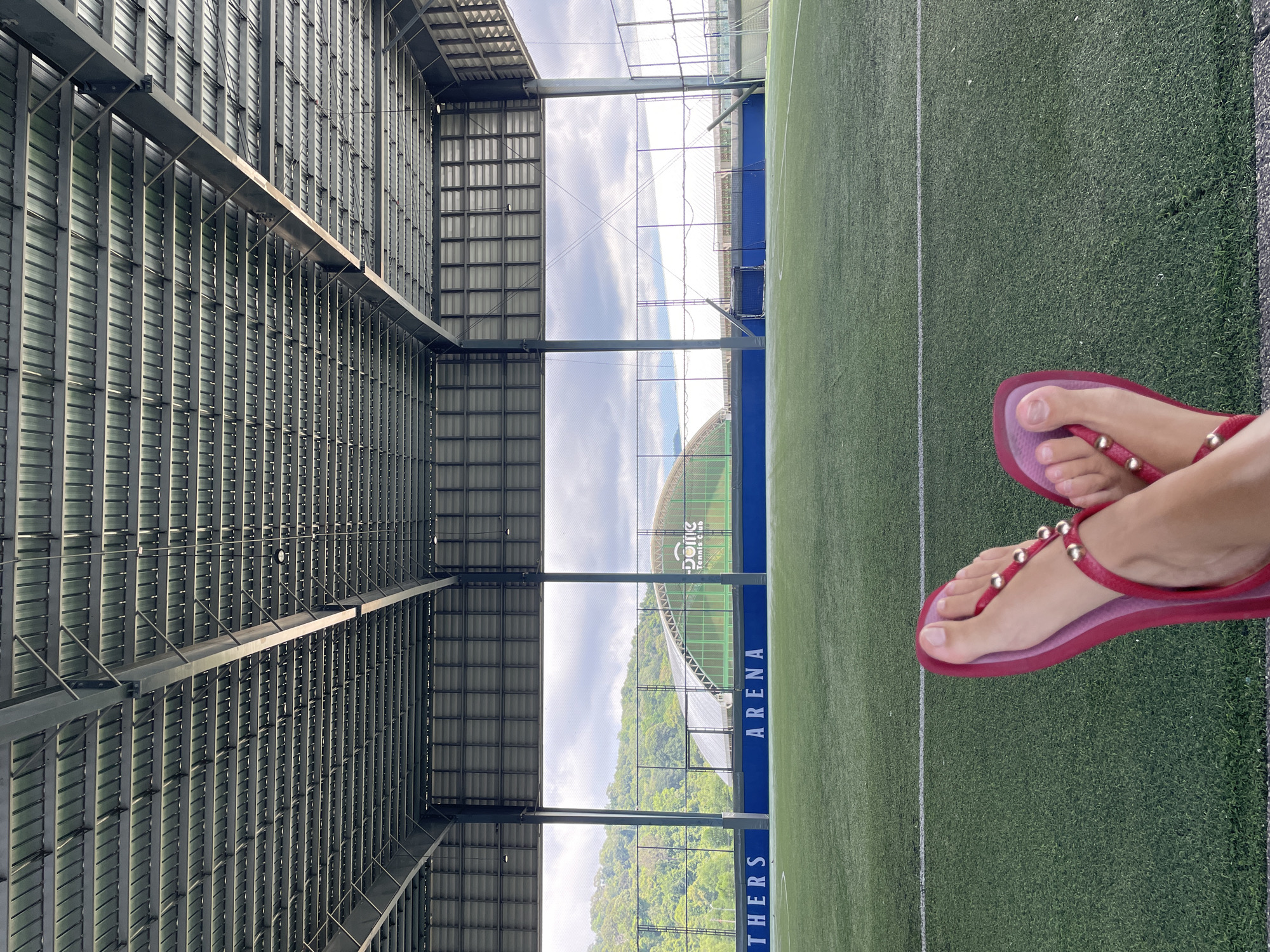 A low-angle, medium shot shows a person's feet, adorned with pink flip-flops, resting on a vibrant green artificial turf soccer field. The feet are crossed at the ankles, with the left foot positioned closer to the camera. The field is marked with white lines and stretches into the distance under a large, industrial-style metal roof. Beyond the field, a mesh fence encloses the area, revealing a lush, tree-covered hillside under a cloudy sky. In the background, a green dome structure with the text "DOME Tennis Club" is visible. The overall scene suggests a recreational facility in a natural setting.