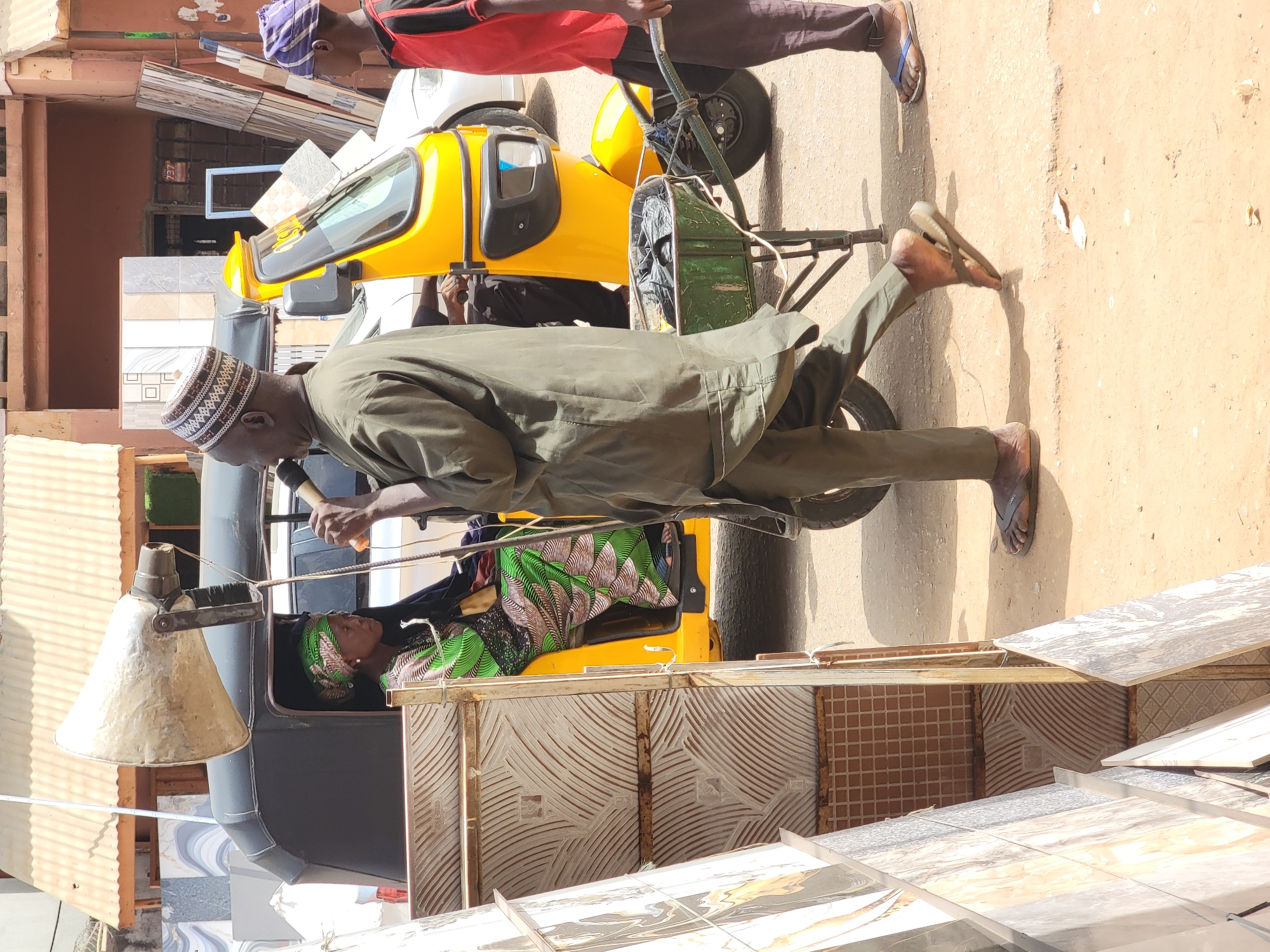 In Maiburiji, Nigeria, a man in a green tunic and patterned kufi propels a yellow and black auto-rickshaw along a dusty street. Inside, a woman wearing a green headwrap lies still. Another man in a red shirt and dark trousers walks in the background. Tiles are stacked along the roadside.