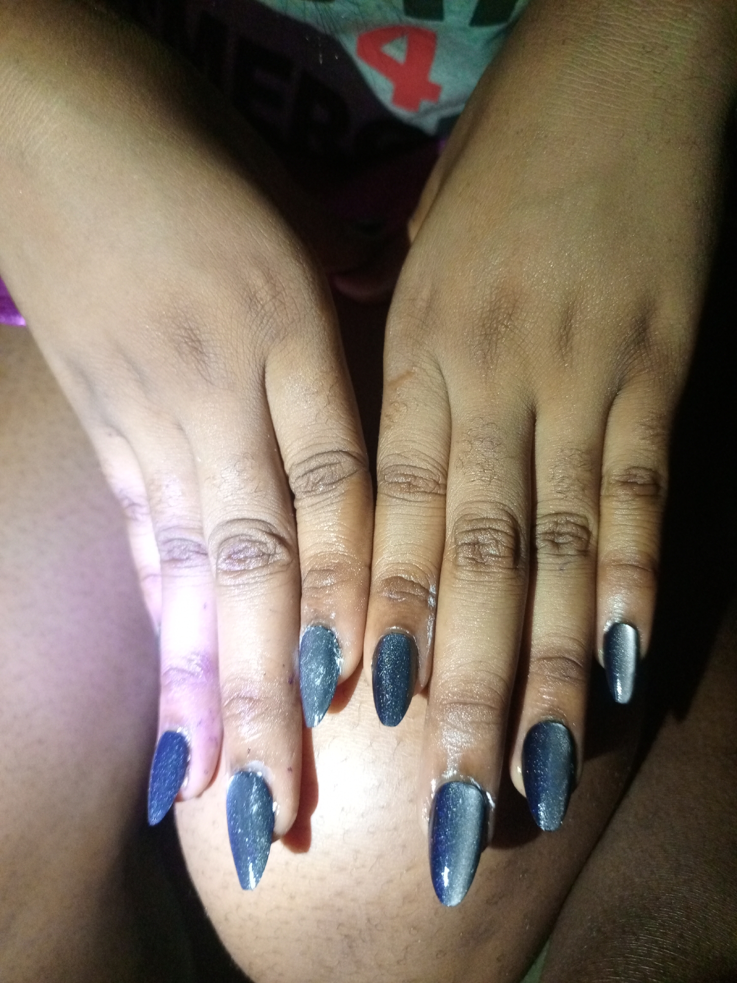 A close-up shot displays two dark-skinned hands with manicured fingernails. The fingernails are almond-shaped and painted with a color-shifting polish that appears blue and grey, with a cat-eye effect. The lighting is uneven, causing shadows and highlights across the hands. The background includes a glimpse of clothing, appearing to be a t-shirt with a visible number. The presence of skin and hair suggests the hands are resting on a leg. Location is not discernible from the image.