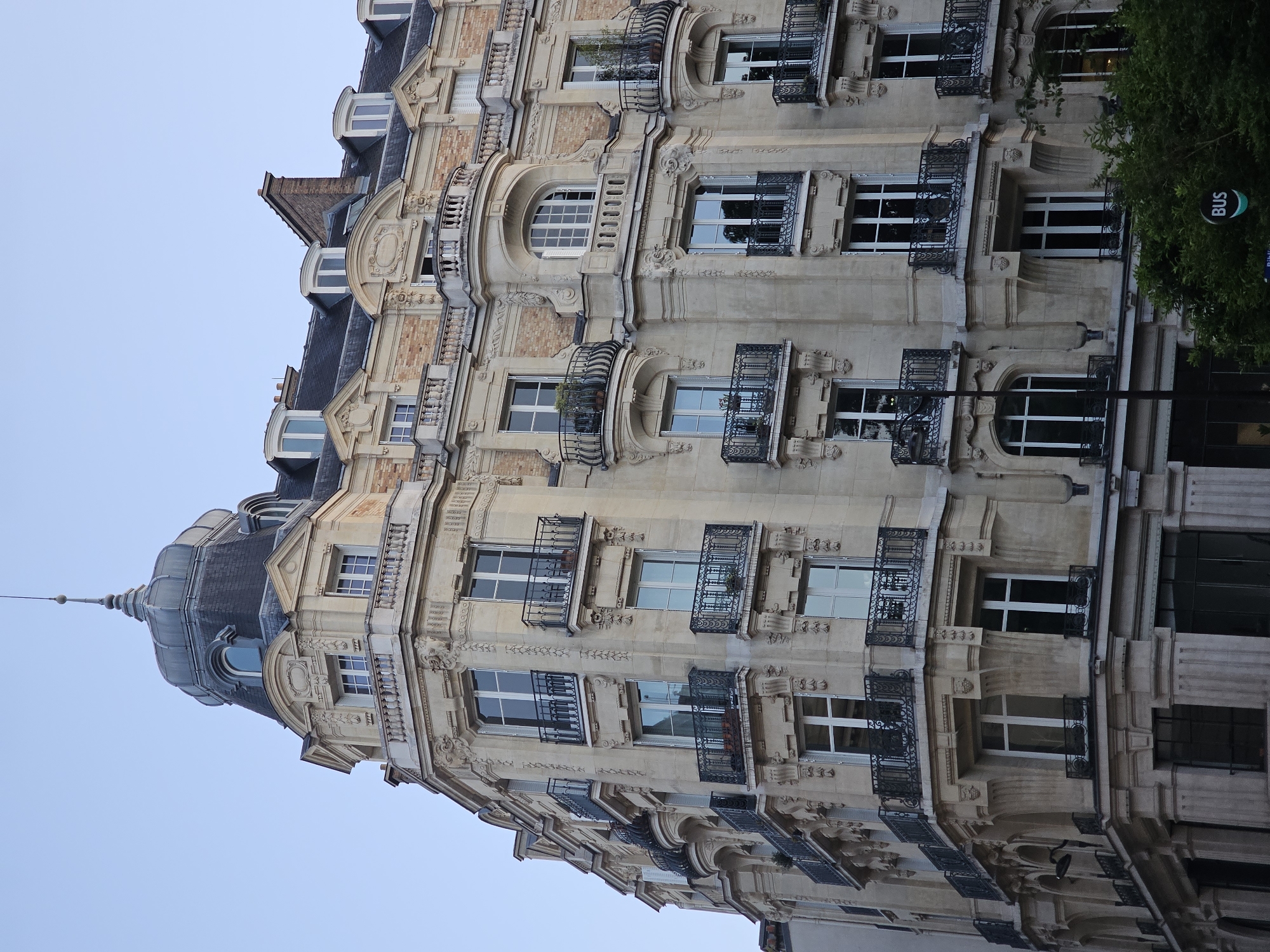 The image shows a building in Paris, France. The building has a domed roof and is decorated with ornate carvings. The windows are rectangular and have black iron railings. There is a "BUS" sign on the right side of the image. The sky is blue.