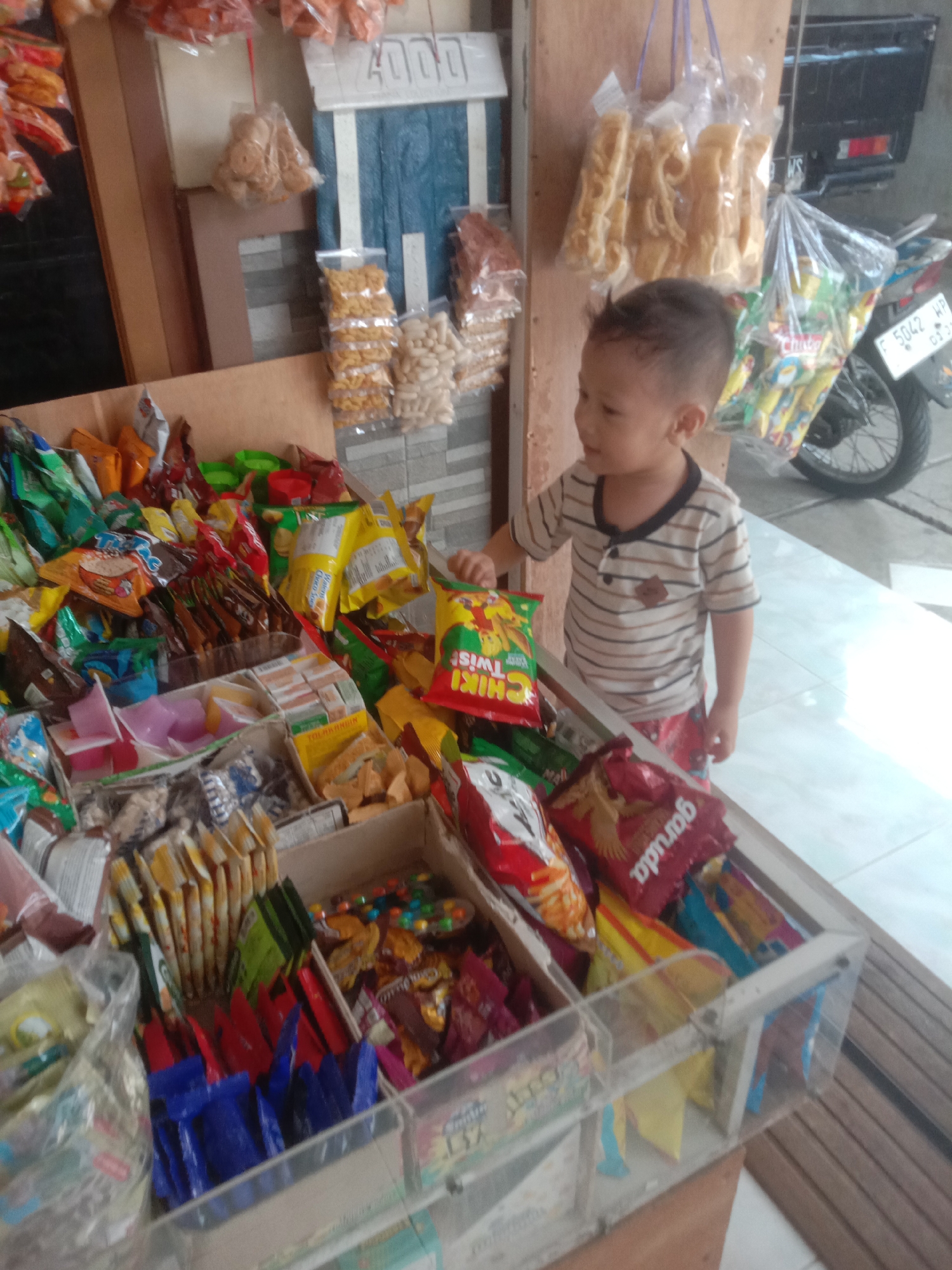 The image depicts a street-side snack stand in Bojong, Indonesia. A young boy, wearing a striped t-shirt and patterned shorts, is examining a bag of snacks. The stand is laden with various packaged snacks, including chips, candies, and biscuits, displayed in transparent plastic bins and hanging bags. Behind the stand, a motorcycle and a black vehicle are visible, along with the exterior of a building with a wooden facade and a tiled floor. The overall scene suggests a typical local marketplace.