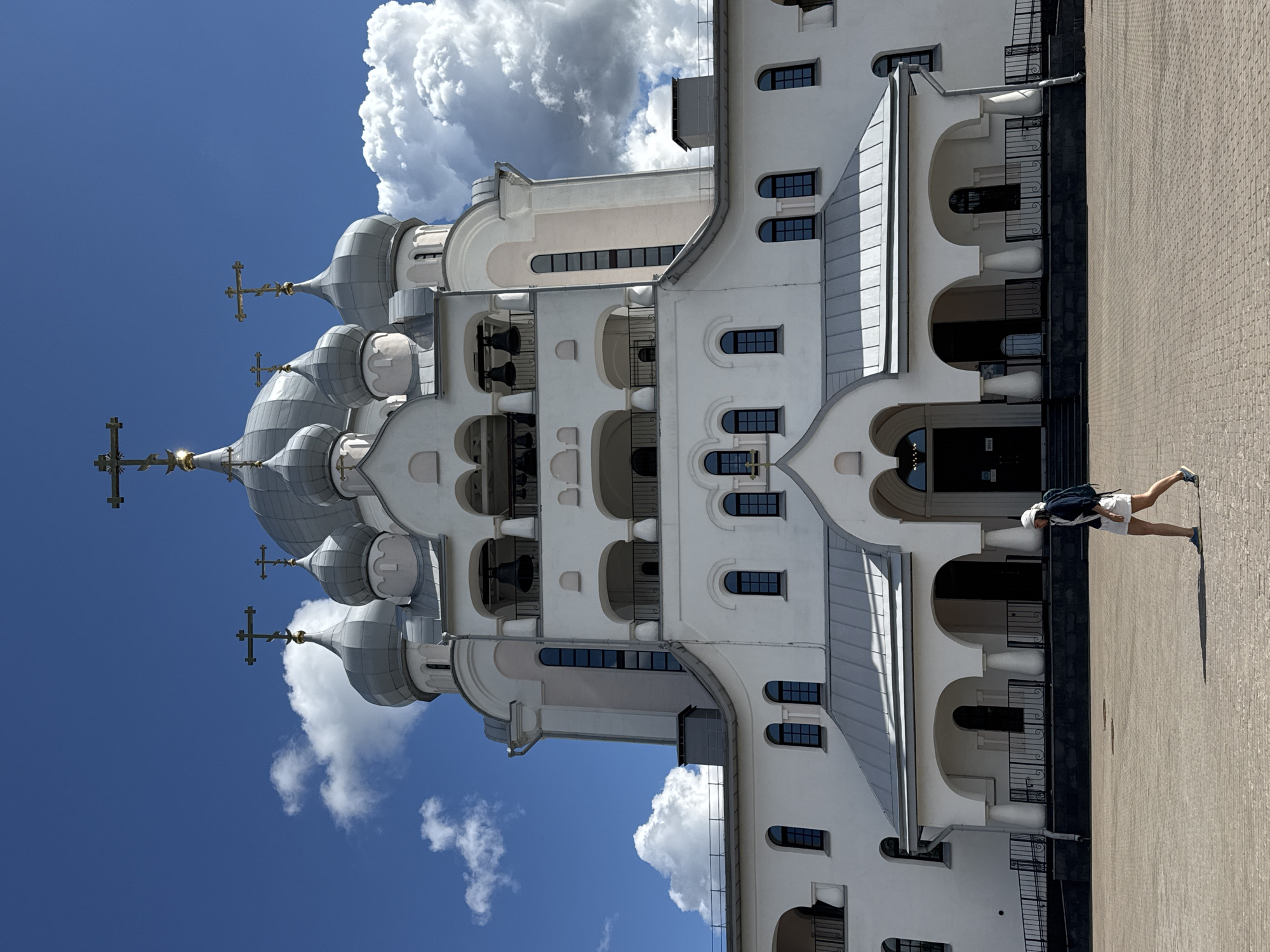 A woman wearing a white cap, a dark jacket, and white shorts walks across a paved square in front of a white orthodox church with silver domes. The church has a large central dome and several smaller domes, each topped with a cross. The building has arched doorways and windows. The sky is blue with fluffy white clouds. The scene appears to be in Saint Petersburg, Russia, likely in the Nevsky District due to the architectural style of the church and the presence of a paved square, which are common features in the city.