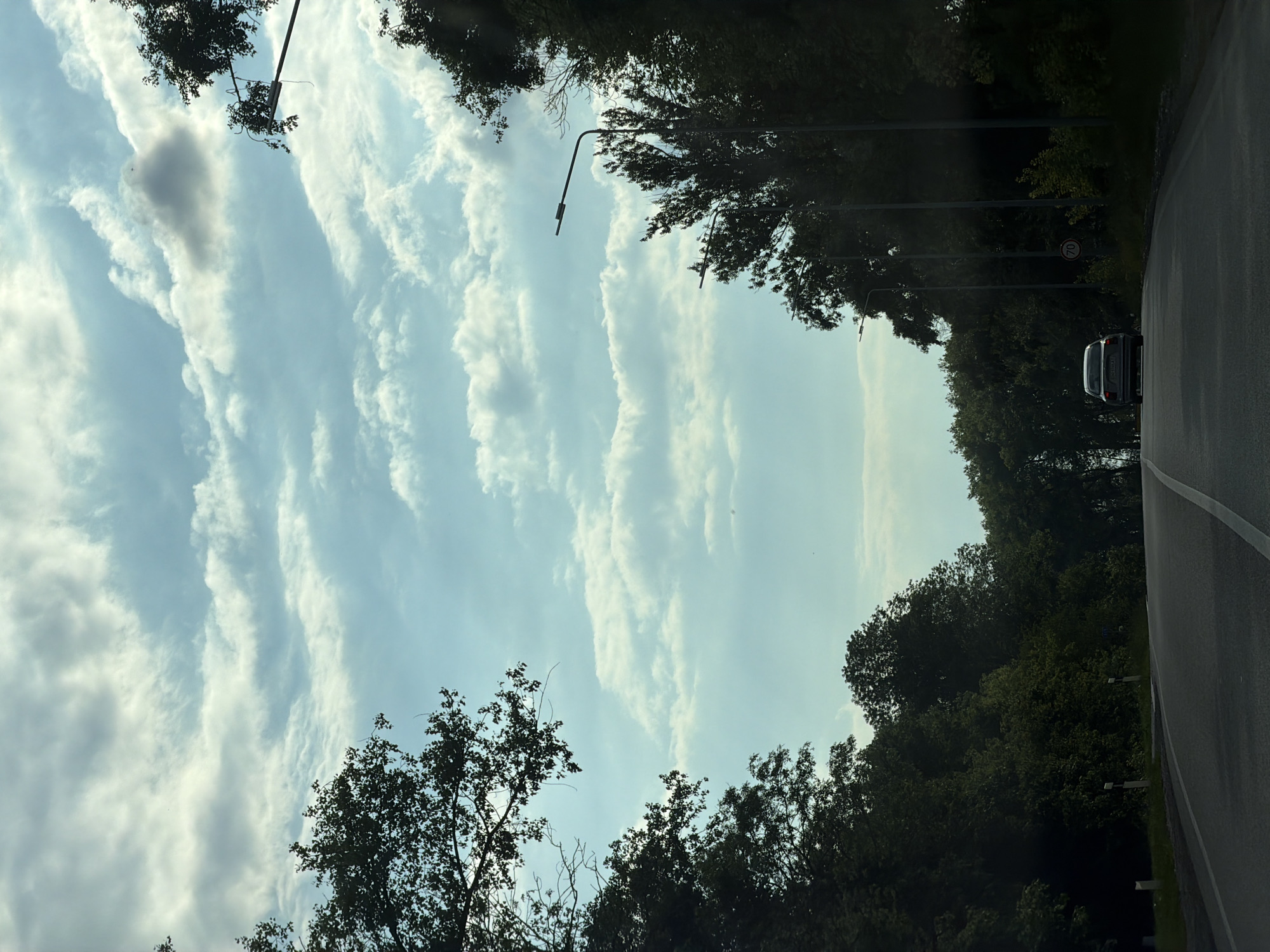 The image depicts a view of a road in Viktorivka, Ukraine, as seen from a vehicle. Tall trees line both sides of the asphalt road, forming a canopy overhead. The sky is visible above the trees, featuring a mix of blue sky and scattered white clouds. A silver car is driving ahead on the road, and several lampposts are positioned along the right side of the road. A speed limit sign indicating "70" is visible on the right side of the road. The lighting suggests it is daytime.