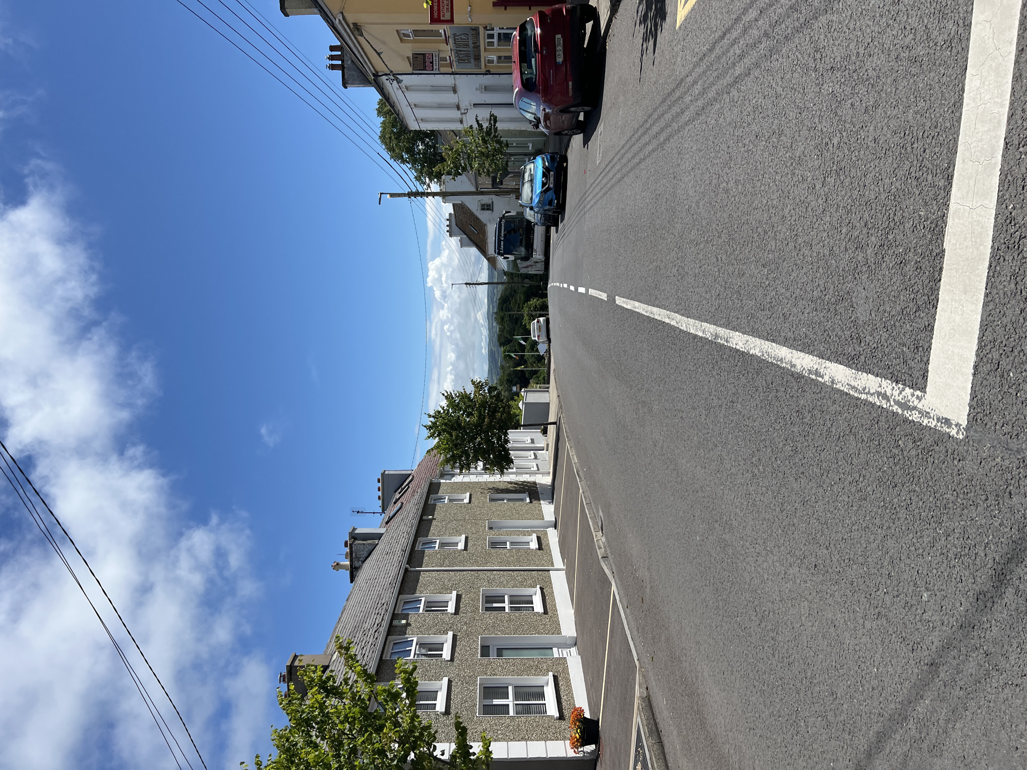 The image captures a view of a street in Mountcharles, Ireland, on a sunny day. The street is paved with asphalt and marked with a white line down the center. On the left side of the street are a row of terraced houses with gray, pebbledash exteriors and white-framed windows. Trees with green foliage are planted in front of these houses.
On the right side of the street, there are also houses, some of which appear to be businesses. One building has a yellow facade and a sign that reads "TASTY BITES." Parked cars are visible along the right side of the street, including a red SUV, a blue hatchback, and a black car. Further down the street, a large white bus with "Liam Connelly" written on its side is parked. In the distance, rolling hills and a hazy landscape can be seen under a bright blue sky with scattered white clouds. Power lines are strung across the sky.