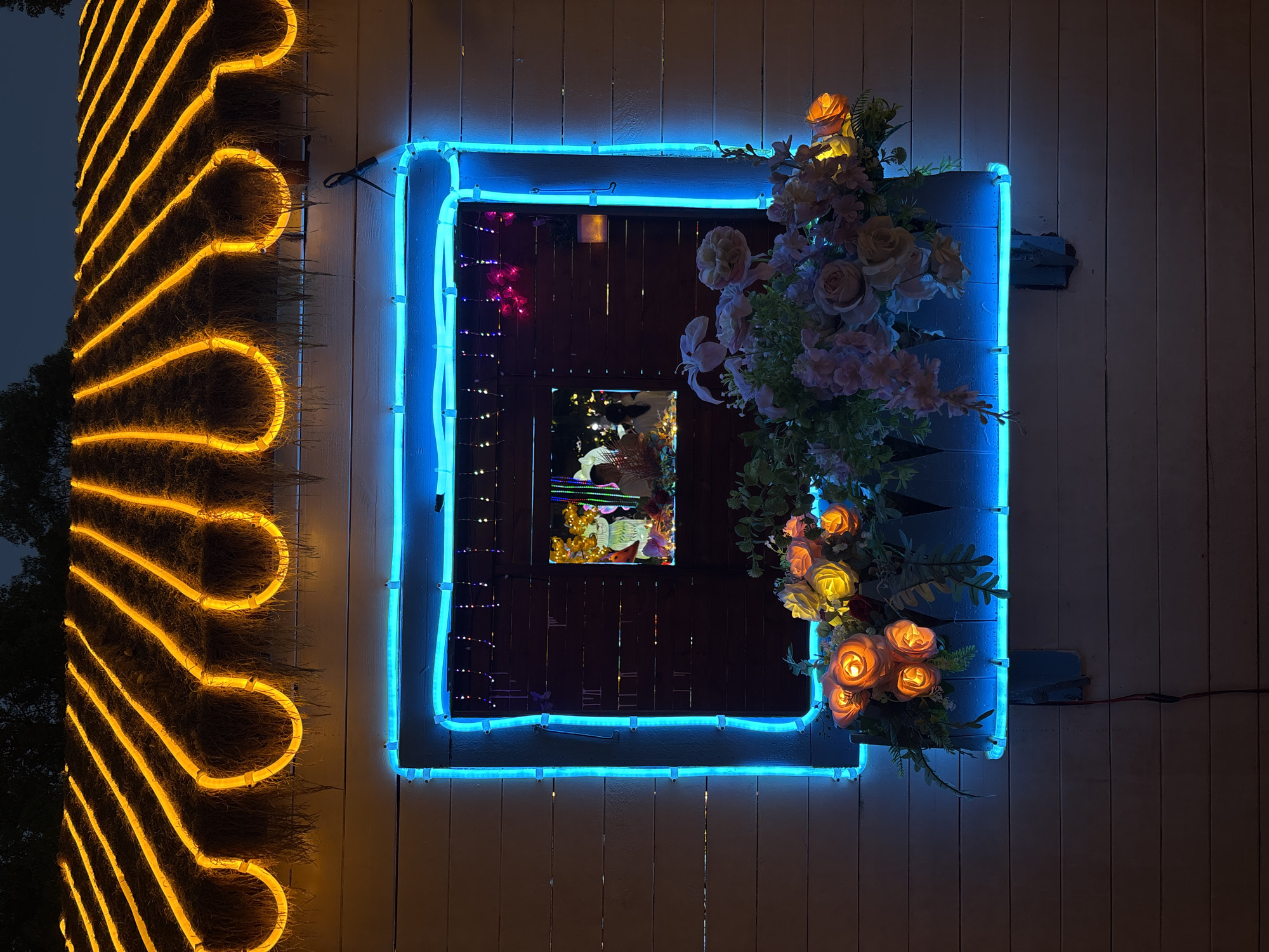 This image shows an outdoor scene at night in Baqiao, China. A white wooden wall is visible, with a window set into it. The window is outlined with bright blue neon lights. Above the window, there is a thatched roof decorated with orange neon lights arranged in parallel, curved lines. Below the window, a blue planter box is filled with artificial flowers. The window itself reflects a scene with people and colorful lights. The overall impression is festive and decorative.
