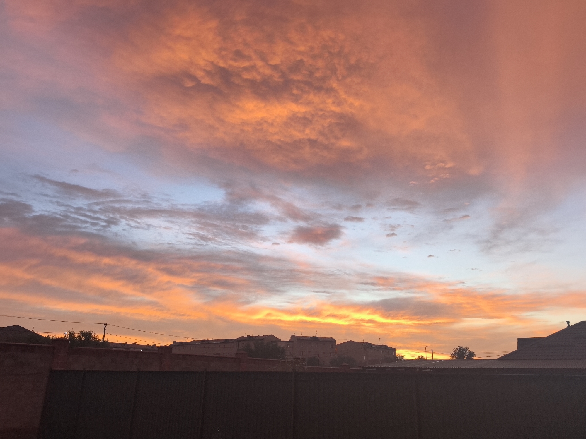 The image depicts a vibrant sunset over a residential area in Taldykorgan, Kazakhstan. The sky is filled with dramatic clouds painted in shades of orange, pink, and purple, transitioning to a pale blue near the horizon. Below the sky, the silhouettes of multi-story buildings and houses are visible, some with visible windows and antennas. A dark fence runs across the foreground, obscuring the lower portion of the scene. Trees are scattered between the buildings. The overall impression is one of a peaceful evening in an urban setting.