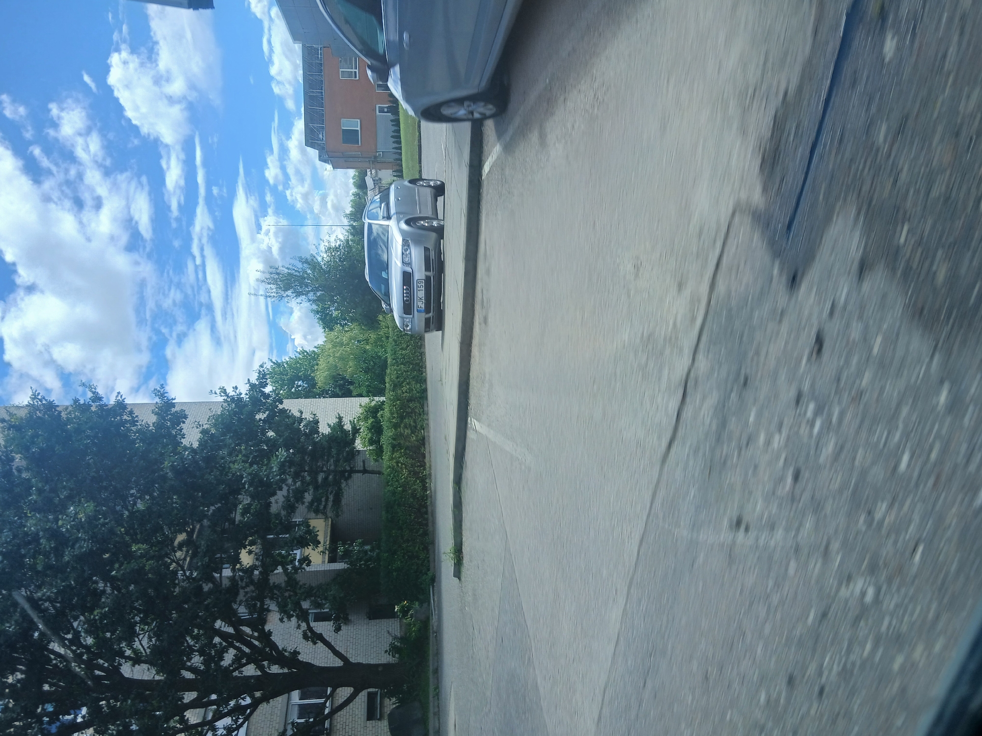 This is a slightly blurry, eye-level, outdoor photograph taken on a sunny day in Garliava, Lithuania. The scene depicts a parking lot with two cars. On the left, a silver Audi A4 Avant is parked. Its license plate reads "FJK 159". To its right, partially visible and out of focus, is the side of another silver car. The background features a light-colored brick building with trees and hedges in front of it, and a modern orange brick building with solar panels on its roof further in the distance. The sky is blue with fluffy white clouds. The foreground is a paved asphalt surface, showing some cracks and a dark rectangular patch, possibly indicating a repair or stain.