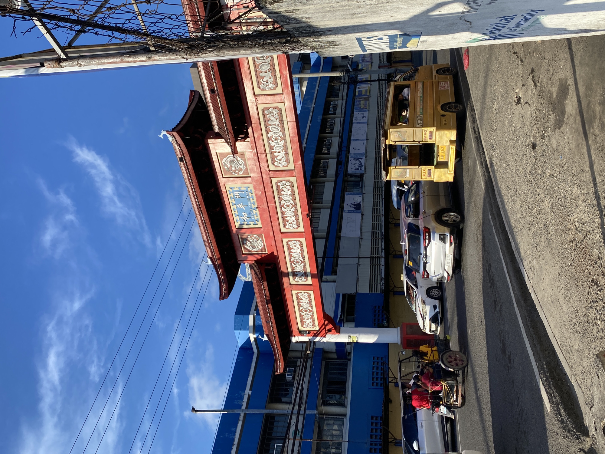This image captures a street scene in Chinatown, Davao City, Philippines, on a bright, sunny day with a clear blue sky and scattered white clouds. The central focus is a large, ornate Chinese-style archway, predominantly red with white decorative panels featuring intricate reliefs of floral patterns and mythical creatures. A blue plaque on the archway reads "和平門" and "PEACE ARCHWAY" in English. A white, stylized mythical creature adorns the apex of the archway's roof.
To the left of the archway, a multi-story building complex, painted primarily in blue and white, features numerous windows and visible air conditioning units. Posters or advertisements are displayed on its lower facade. Overhead power lines stretch across the upper left portion of the frame. To the right, a concrete wall topped with a chain-link fence runs vertically, partially obscuring the view. A blue sign attached to the wall partially displays "DAVIES" and "Habitat for Humanity".
On the asphalt road, a moderate amount of traffic is present. In the immediate foreground, a custom-built, open-air tricycle with a red-wheeled chassis and a black canopy is visible, carrying at least three passengers: two in red shirts and one in a yellow shirt. Behind it, a silver SUV (Toyota Raize, license plate AAS 1197) is stationary. To its right, a white sedan (Toyota Vios), identifiable as a taxi by a yellow sign on its roof, is also stopped. Further right, a yellow multi-cab or jeepney-style public utility vehicle (license plate AAF 8345) is seen from the rear, displaying "KEEP DISTANCE," "MATINA PRK. 10," and "DAVAO" on its tailgate. The road surface shows some cracks and patches. Shadows cast across the road and the concrete barrier on the right indicate a low sun angle.