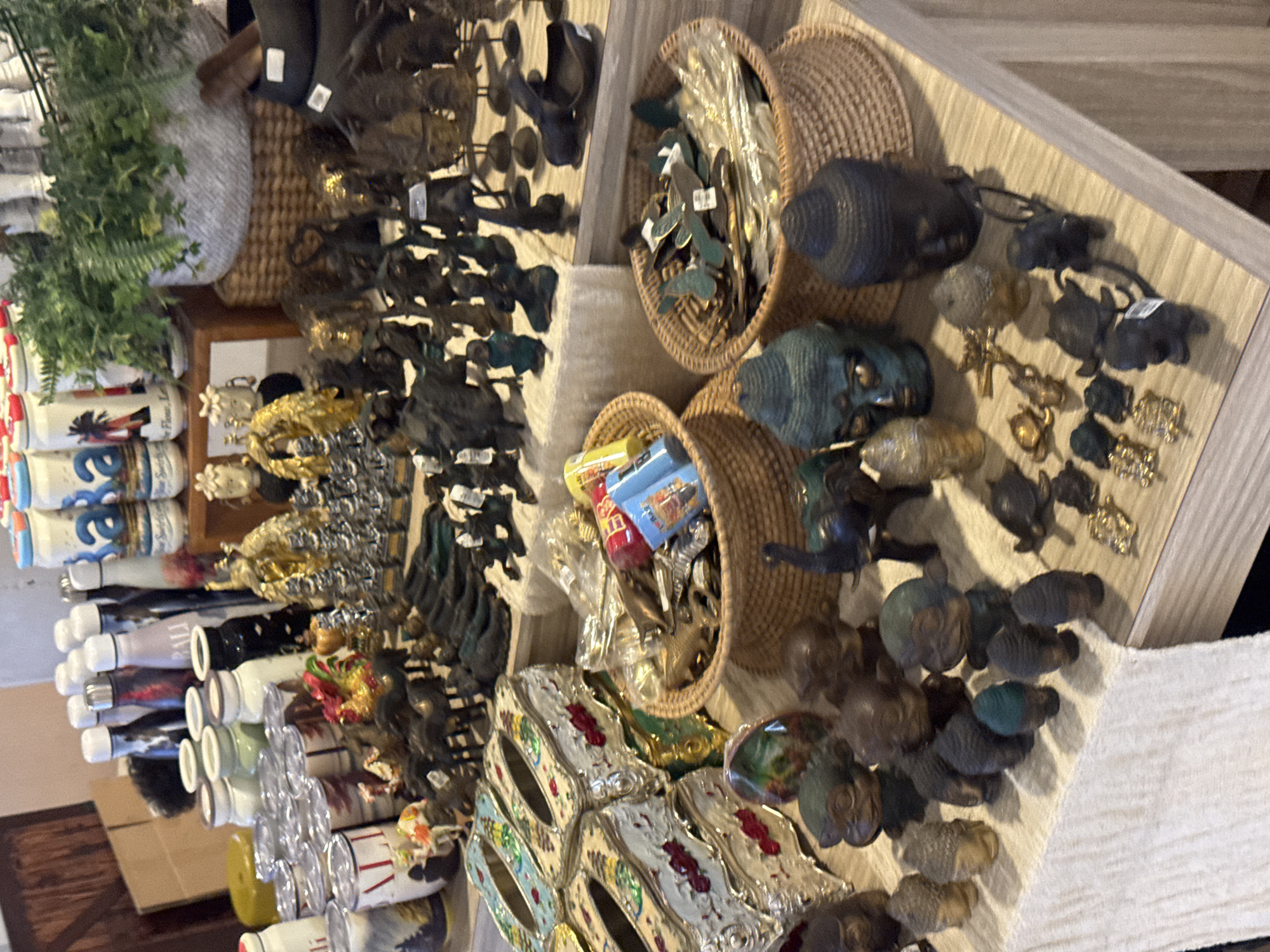 The image shows a retail display of decorative items, likely in a gift shop. The products include an assortment of small statues, figurines, and decorative containers. In the foreground, there are several woven baskets filled with small metal figurines, including butterflies and what appear to be small animals. There are also several Buddha head sculptures and monkey figurines. To the left, there are decorative tissue box covers. In the background, there are various bottles and canisters, some with colorful labels. A potted fern is visible on a wooden shelf in the upper right. The overall impression is of a souvenir or craft shop. Given the context, this shop is likely located in Kuta, Indonesia, a popular tourist destination known for its markets and gift shops selling similar items. There are no people visible in the image.