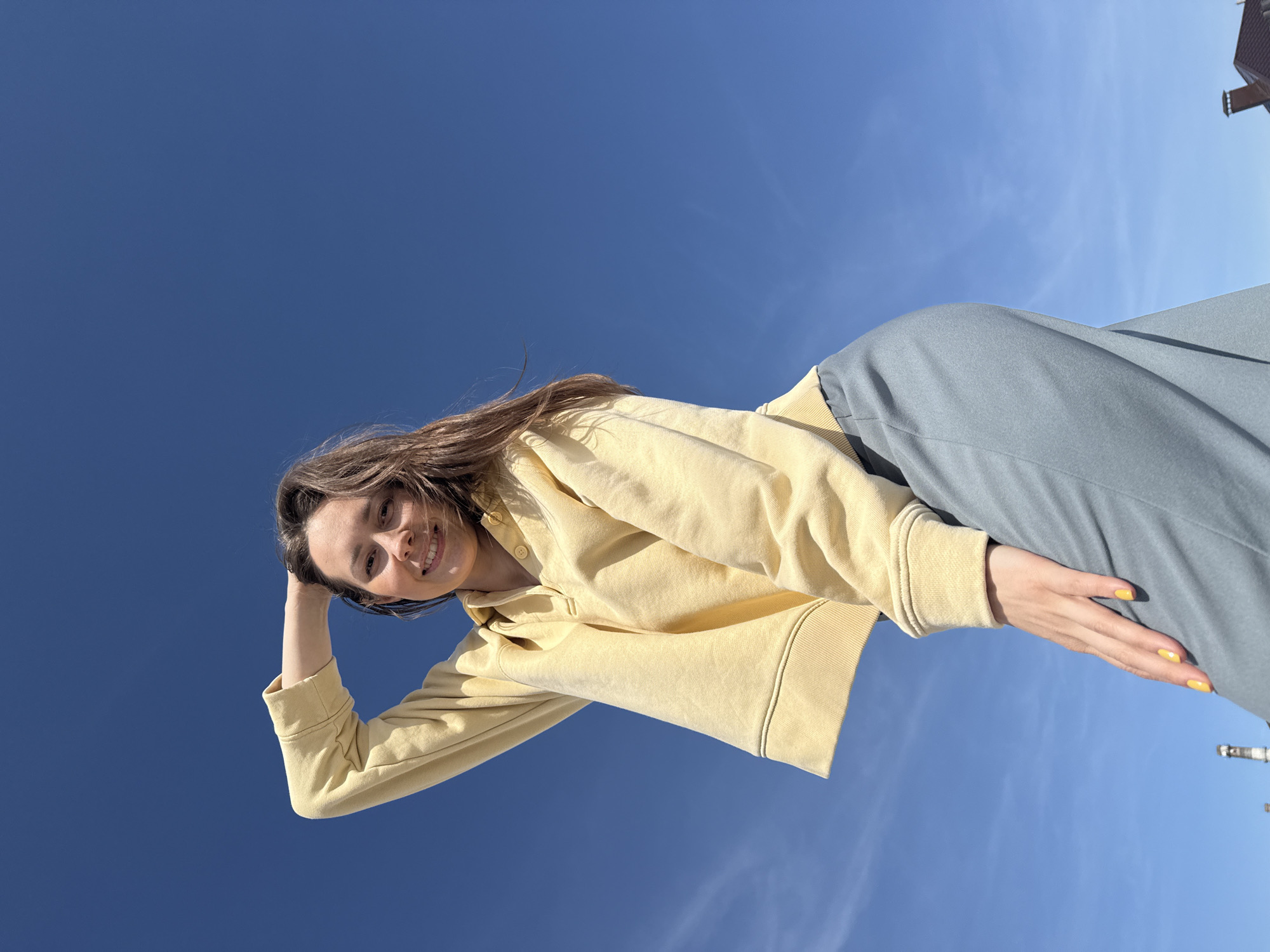 A young woman is viewed from a low angle against a blue sky. She is wearing a yellow cropped sweatshirt and grey-blue pants. Her right hand is resting on her head, and her left hand is resting on her leg. She is smiling. In the far lower right corner, there are architectural details of an industrial structure, which suggests an urban location. This is Chaikovsky, Russia.