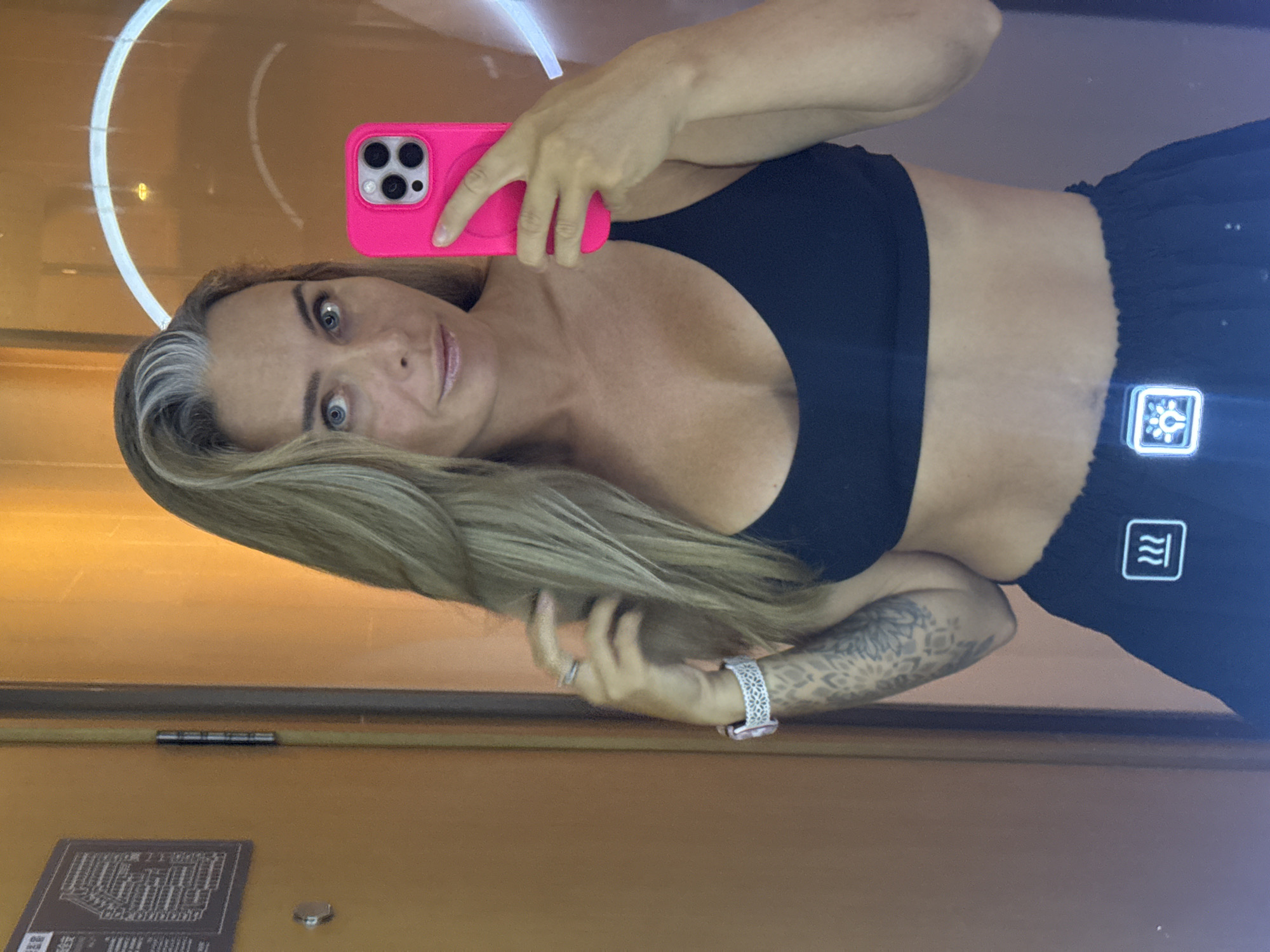The image is a selfie taken by a woman with blonde hair and blue eyes. She is wearing a black sports bra and black pants. She is holding a pink smartphone in her right hand. She has a tattoo on her left arm and is wearing a watch on her left wrist. The background appears to be a room with orange walls. There is a door on the left side of the image with a diagram on it. The woman is in a selfie pose, looking at the camera. There is no discernible indication of the location being in the city of Shanghai, China.