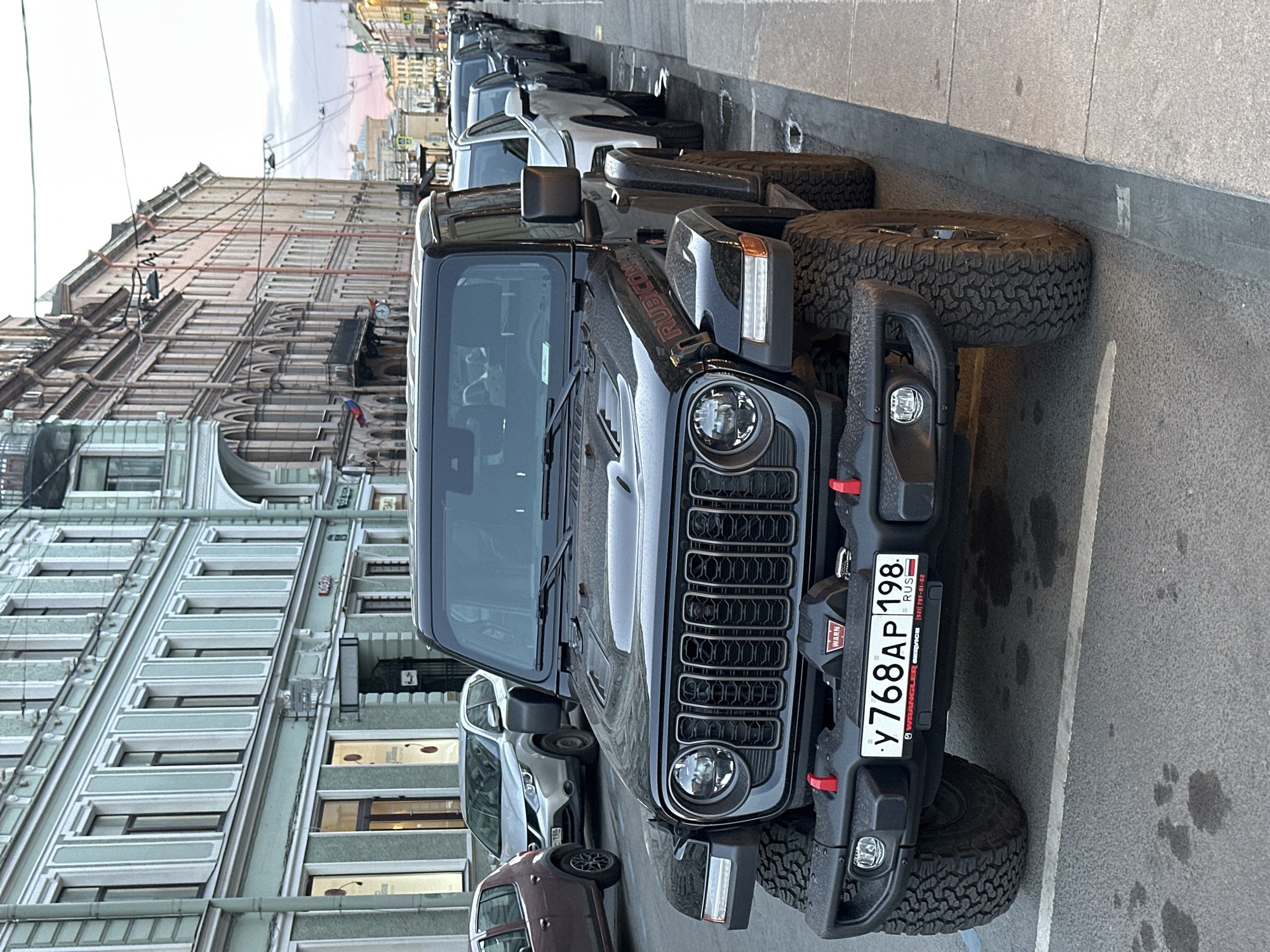 The image depicts a black Jeep Wrangler Rubicon parked on a street in Saint Petersburg, Russia. The license plate reads "У 768 АР 198 RUS." The Jeep is parked alongside other vehicles. The buildings lining the street are characterized by European-style architecture. The sky is overcast. The street appears to be asphalt, with markings indicating parking spaces.
