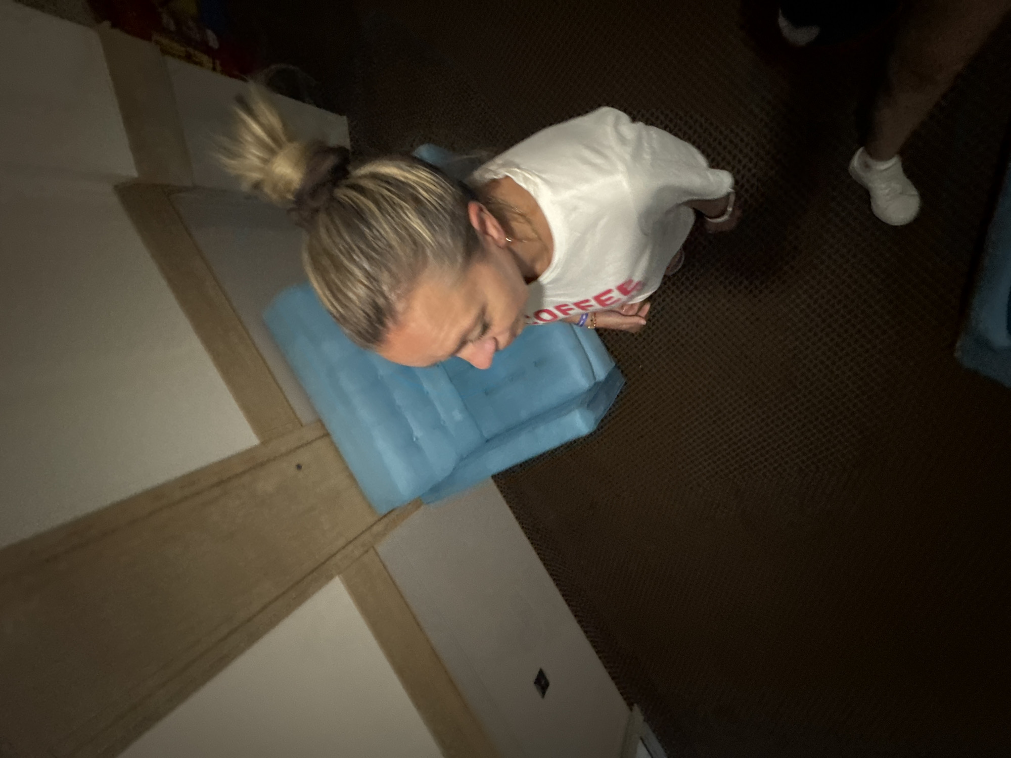 A high-angle medium shot captures a blonde woman with her hair in a messy bun, wearing a white long-sleeved shirt with red lettering that appears to say "COFFEE" down the front. She is standing and looking to her left, slightly down. The shirt has some red lettering visible. She wears a bracelet on her left wrist. In the background, there is a small, bright blue chair with a tufted surface. The wall behind the chair is painted a light color and has a wooden beam running horizontally across it, with another beam above it. The floor is carpeted in a dark brown, textured material. To the right of the woman, a pair of white sneakers is visible, suggesting another person is present but out of frame. The scene appears to be indoors, possibly in a hotel room or a similarly furnished space. There is no discernible indication of the city of Nesebar, Bulgaria.
