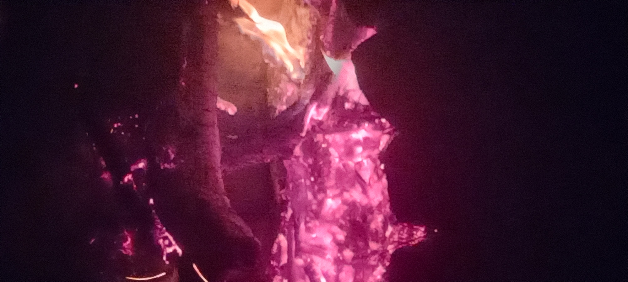The image displays a nighttime scene dominated by a fire. A large bed of intensely glowing pink and purple embers occupies the lower portion and extends towards the center-left. Above this radiant mass, dark, charred wood or logs are visible. To the right, brighter orange and yellow flames rise, indicating an active combustion area. The immediate environment beyond the fire is obscured by darkness, with no discernible background features or structures. No individuals, specific objects beyond the fire's fuel, or discernible actions are present within the frame. The location within the city of Kryvyi Rih, Ukraine, cannot be determined from the visual information provided in the image.