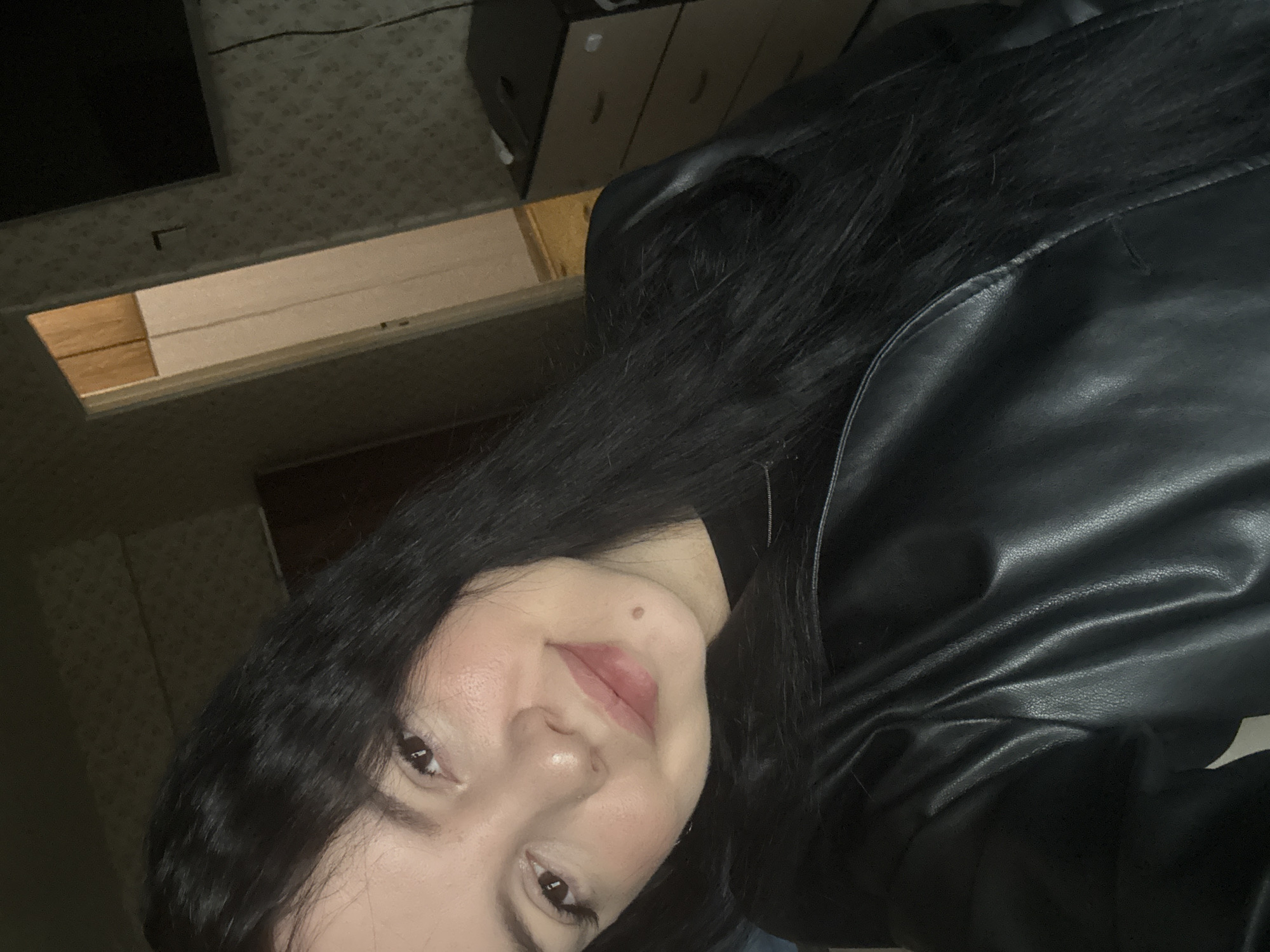 A close-up, rotated portrait shows a woman with long, dark hair wearing a black leather jacket. Her face is angled towards the camera, revealing fair skin, dark eyes, and a mole on her upper lip. The background includes a patterned wall, a dark television screen, a wooden shelf or drawer partially pulled out, and a light-colored set of drawers with curved handles. The location cannot be determined to be in Mytishchi, Russia based on this image.