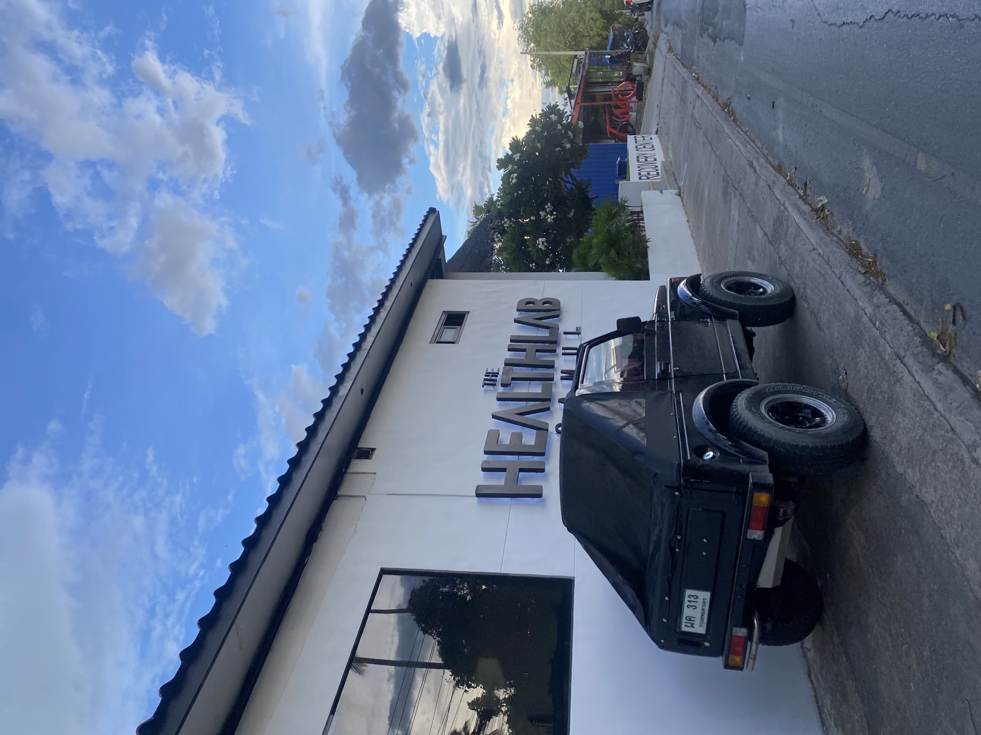 A ground-level, outdoor shot captures a black, four-wheeled vehicle parked alongside a light gray sidewalk and adjacent to a white, single-story building in บ้านบางรักษ์, Thailand. The building’s facade features dark gray signage reading "THE HEALTHLAB INTL" and a dark-framed window reflecting trees and sky. The vehicle, oriented with its rear towards the viewer, displays a license plate reading "ผล 313" in Thai script. The background reveals additional buildings and lush trees, situated beneath a partly cloudy sky. The lighting suggests either early morning or late afternoon.