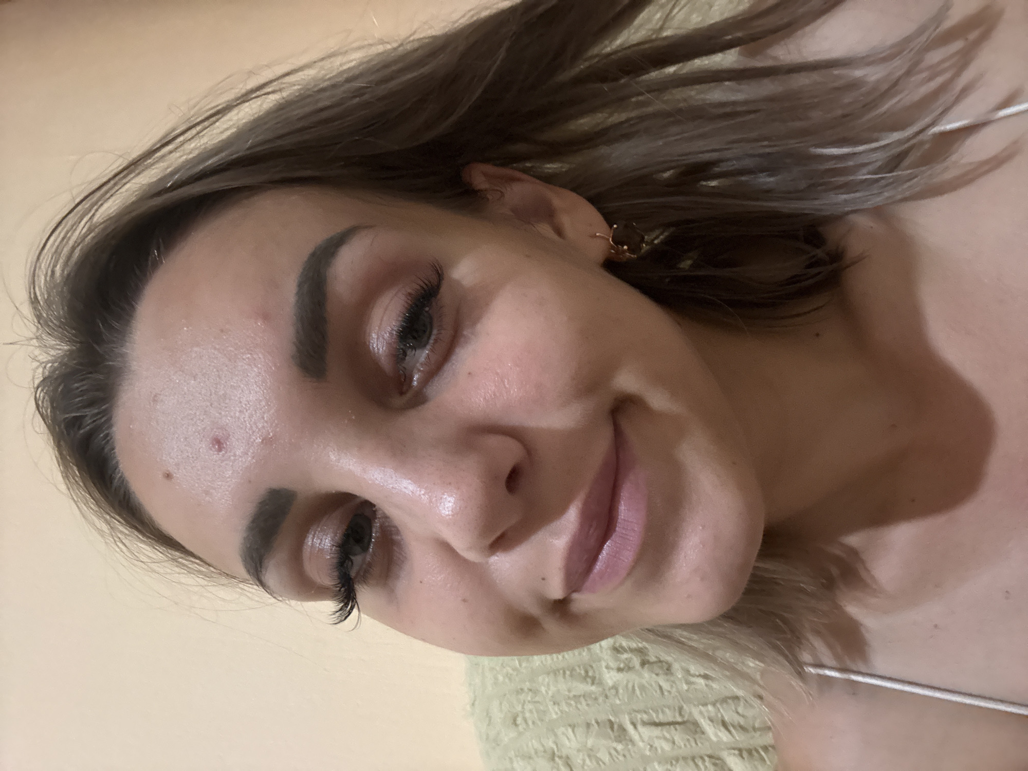 The image is a close-up, selfie-style photograph of a young woman. She has light brown hair that is slightly windswept and falls around her shoulders. Her eyebrows are dark and well-defined, and she is wearing noticeable false eyelashes. Her eyes are open, and she is looking towards the camera. She has a faint smile, with her lips slightly parted. There are a few small blemishes visible on her forehead and chin. She is wearing a simple white strap top and a small, dark-colored earring in her visible ear. The background is a plain, light-colored wall, with a hint of a textured, light green fabric on the left side of the frame. The lighting appears to be from a direct source, causing some shine on her skin. No specific location in Kholmsk, Russia is identifiable in this image.