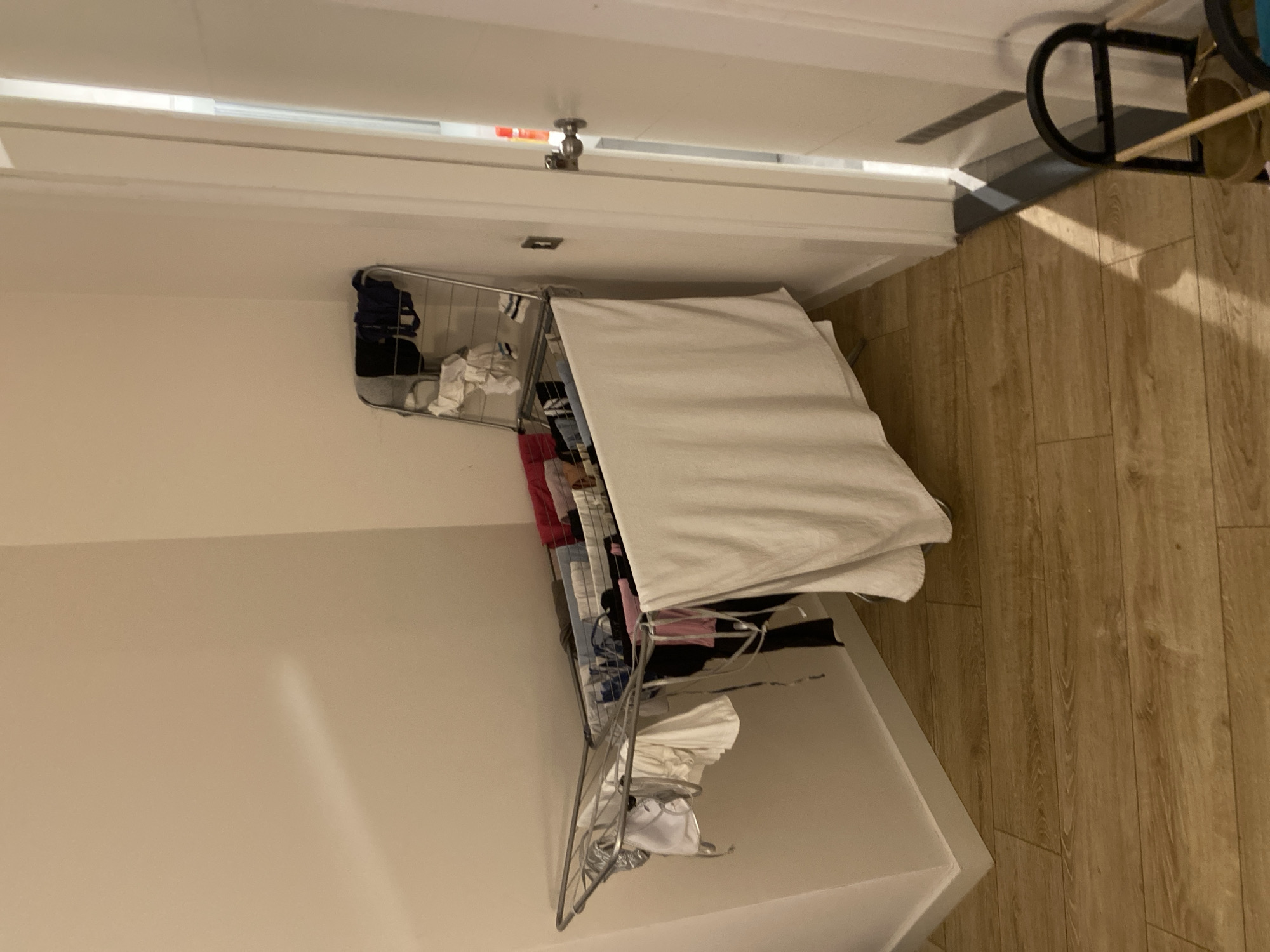 An interior shot shows a corner of a room with a clothes drying rack. The drying rack is silver and metal, and it is filled with various items of laundry, including white towels, dark-colored clothing, and some brightly colored items. A large white towel is draped over the front of the rack. The walls are painted a neutral beige color, and the floor is covered in light-colored wooden planks. To the right of the drying rack is a white door, slightly ajar, revealing a glimpse of another room. Sunlight streams through the doorway, casting shadows on the floor and wall. The overall scene suggests a domestic setting, likely a laundry room or a bedroom. There is no indication of the city of Aksu, Turkey, in the image.