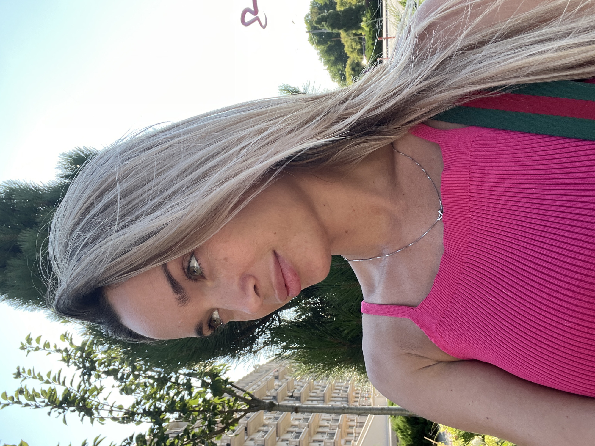A medium shot captures a woman in a bright pink top standing outdoors. She has long, blonde hair and is looking to her left. Her face is visible from the side and features freckles. She is wearing a thin silver necklace. The background includes greenery, a building with multiple balconies, and a clear sky. The image appears to be taken in the city of Fontanka, Ukraine, based on the architectural style and vegetation.