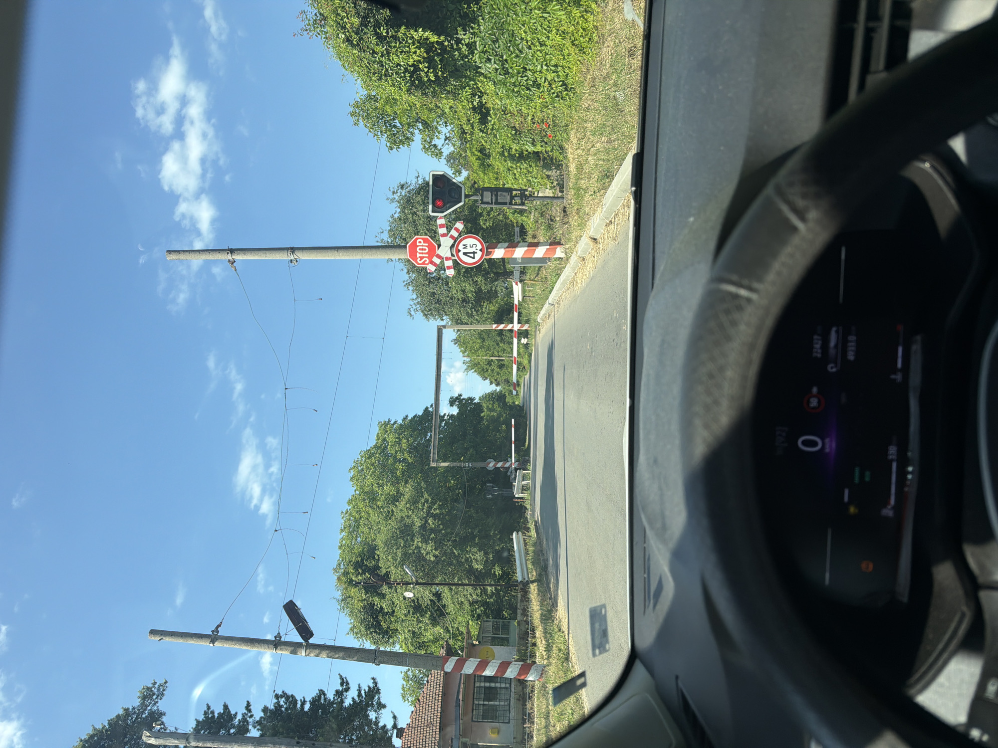 The image depicts a view from inside a vehicle approaching a railway crossing in Borovo, Bulgaria. The crossing is equipped with red and white striped barrier posts, a STOP sign, a red traffic light indicating a stop, and a circular sign with "4.5 M" indicating a height restriction. Two white horizontal barriers are lowered across the road, blocking traffic. The sky is blue with scattered white clouds. Trees and vegetation are visible on both sides of the road, with a small building to the left. The vehicle's dashboard is partially visible, showing a digital display with speed and other indicators.