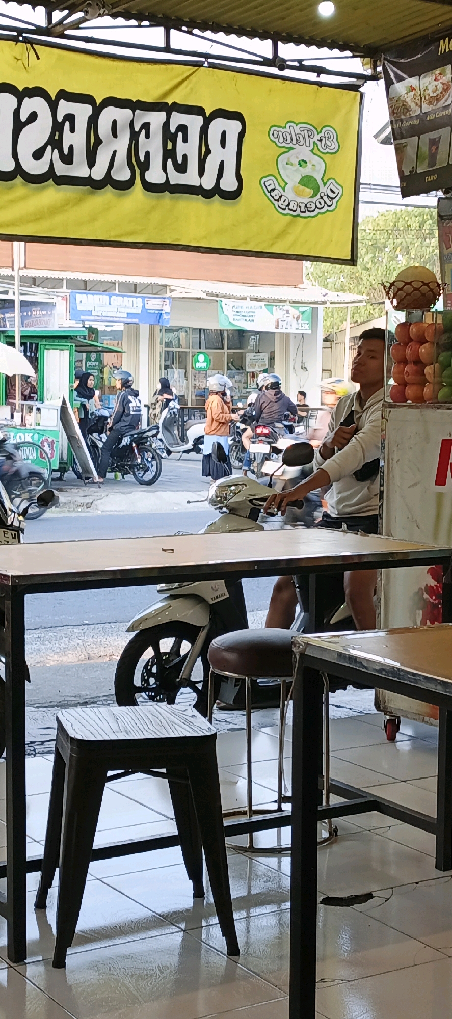 This image captures a street scene in Sidoarum, Indonesia, likely near a food or drink vendor. In the foreground, a wooden-topped table with a metal frame is visible, with a dark stool to its left and another stool to its right. Beyond the table, a white scooter is parked.
The background reveals a bustling environment with several people on motorcycles, suggesting a busy street. A large yellow banner with white text, possibly advertising a business, hangs above the scene. The text on the banner appears to be reversed, reading "REFRES" from right to left. To the right of the banner is a smaller graphic with green elements and text.
Further back, several buildings are visible, including what appears to be a small shop or stall with a green facade and an awning. People are seen standing and interacting, some wearing helmets. A stack of colorful fruits, possibly apples and possibly limes or small melons, is displayed in a basket on the right side of the image. The ground is tiled and slightly wet in places, with visible cracks in the tiles near the table leg on the far right. The lighting suggests it is daytime, with sunlight casting shadows.
