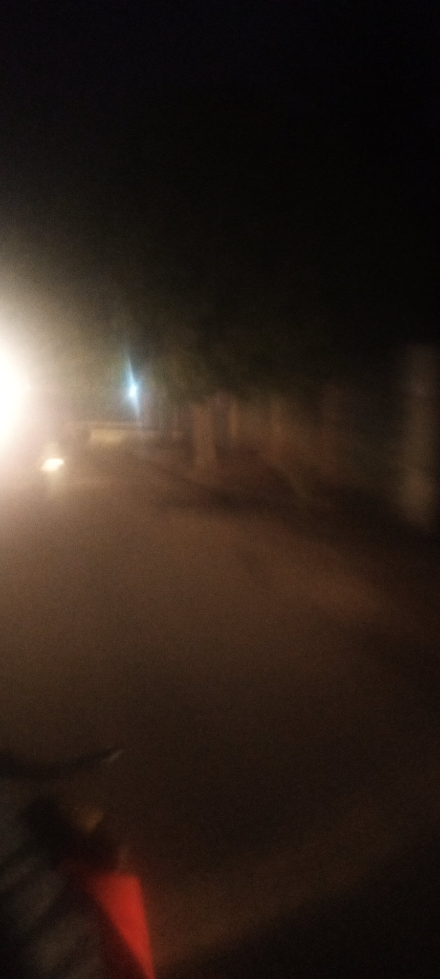 The image shows a blurry street scene at night in Narayi, Nigeria. There is a bright light source on the left side of the frame. A dim street lamp is visible in the distance. Along the side of the road, there are trees and what appear to be buildings or a fence. A part of a bicycle is visible in the bottom left. The overall scene is dark.