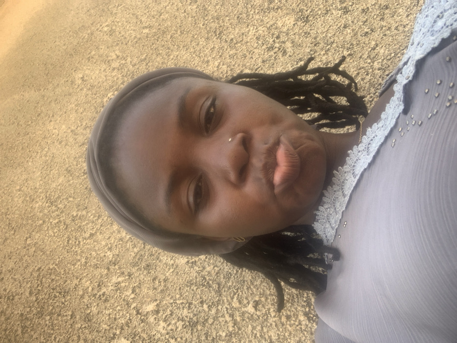 A close-up image taken in Bojude, Nigeria, depicts an adult female positioned against a textured, light brown or tan wall. The female has dark skin, dark dreadlocks partially visible from beneath a plain medium grey headwrap, and a small silver-colored nose stud on her left nostril. Gold-colored hoop earrings are visible in her ears. She is wearing a light grey garment with a light blue or grey lace trim at the neckline, adorned with small silver-colored embellishments. Her lips are pursed, creating a "fish face" or "duck face" expression, and her gaze is directed towards the camera. The scene is illuminated by natural outdoor light.