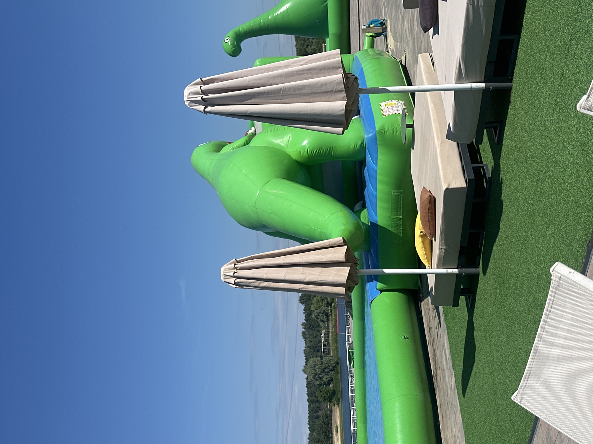 The image shows an outdoor scene at a water park with inflatable dinosaur structures in a lake in Khotianivka, Ukraine. In the foreground, there are several lounge chairs covered with tan cushions and pillows. Two closed beach umbrellas are positioned between the lounge chairs. The ground is covered with artificial green turf and a paved area. In the background, large green inflatable dinosaurs are set up in a blue water pool. The sky is clear and blue, and a treeline with a body of water is visible in the distance.