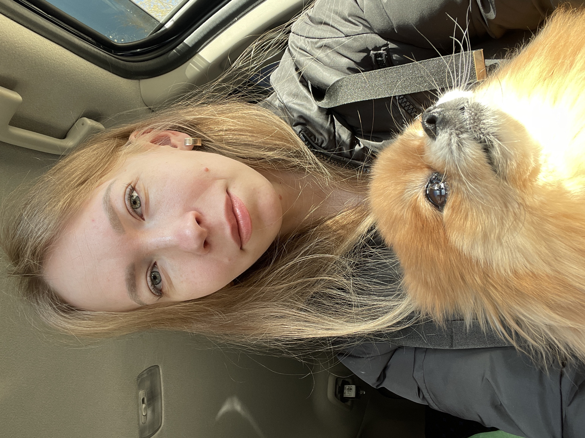 A woman with light blond hair and fair skin is in the passenger seat of a car, wearing a black jacket and seatbelt. She is positioned close to the camera, facing the viewer with a slight smile. Next to her, a small, light-brown Pomeranian dog is looking up at her. The interior of the car is visible, including part of the car roof, a window, and the car's interior upholstery. The image appears to be taken from a slightly rotated angle. The exact location within the city of Perm, Russia is not definitively identifiable from the image.