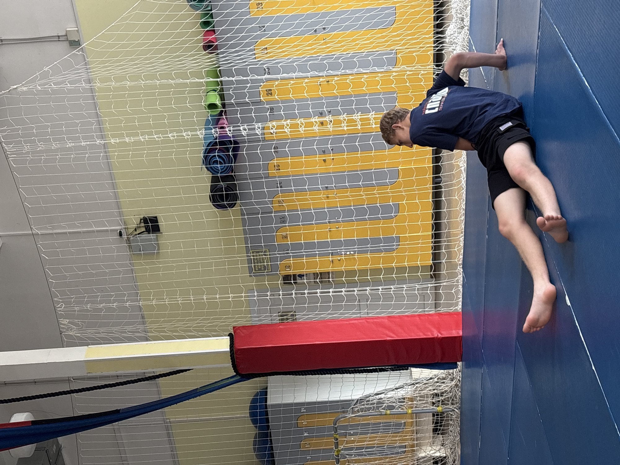 The media file shows a young person in a gymnasium. The person, wearing a dark blue t-shirt and black shorts, is upside down, with their feet on the blue mat and their hands on a yellow and grey wall. The gymnasium features a high ceiling with bright yellow and grey panels on the wall. A white net stretches across the upper part of the scene, and a red padded mat is visible along the bottom edge. Several rolled-up mats in various colors are stacked on the left side of the image. The overall impression is of a gymnastics or acrobatic training environment. The lighting suggests an indoor setting, likely during the daytime. There is no discernible text in the image.